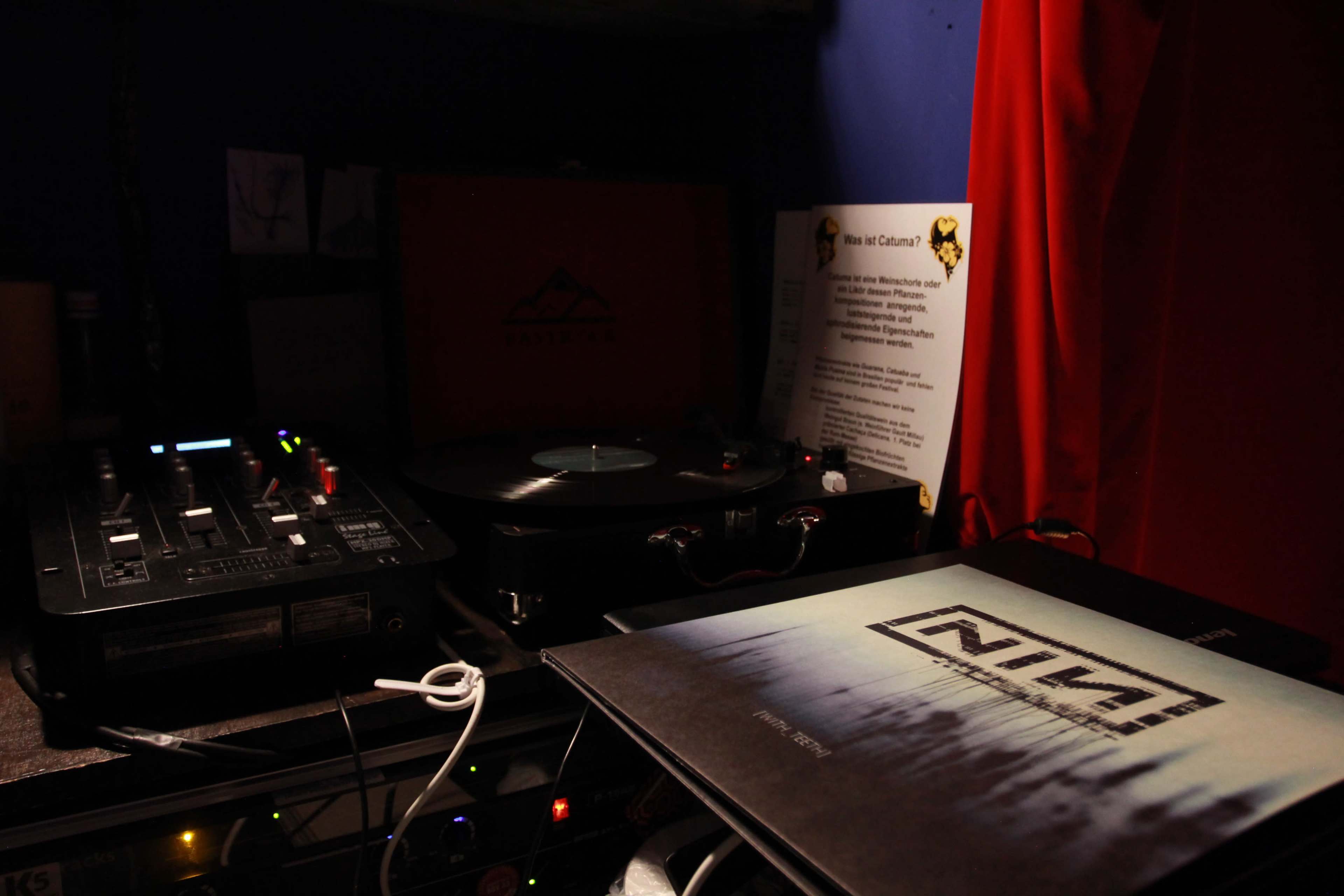 A turntable and mixer are set up on a table beside a vinyl record and informational flyer in a dimly lit room.