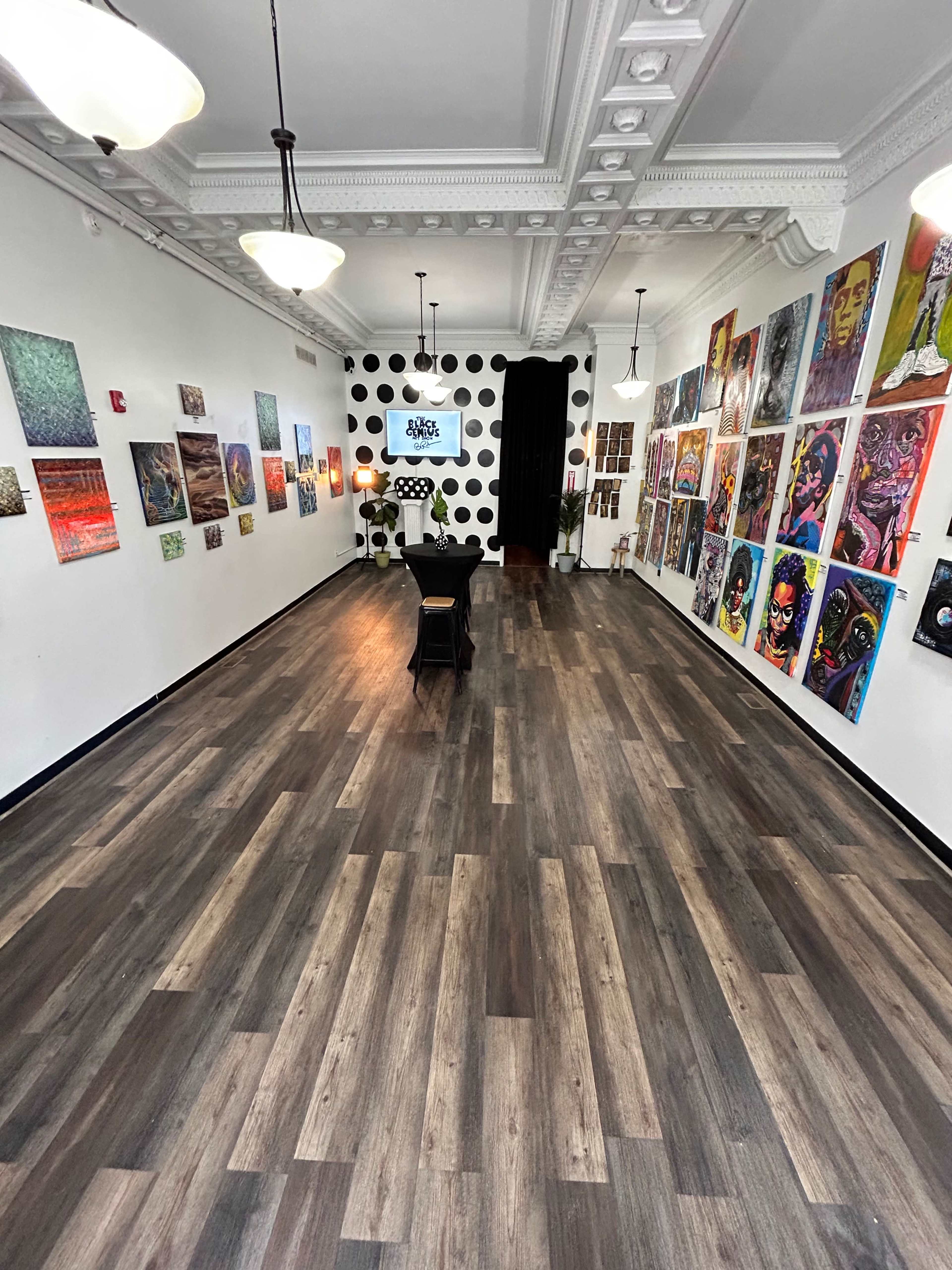 An art gallery with wooden flooring, displaying a variety of colorful artwork on the walls and featuring furniture in the center of the space.