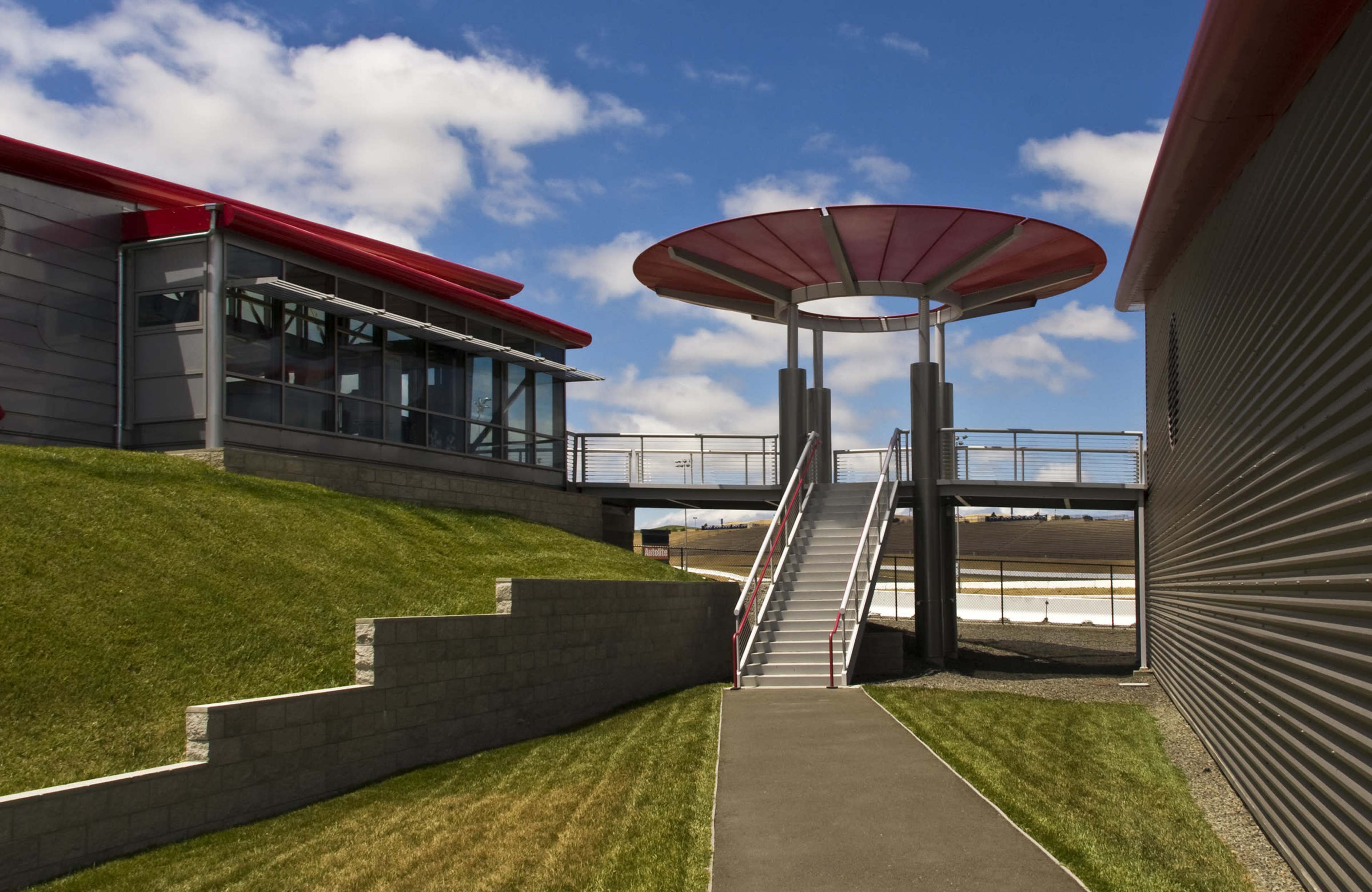 A modern building with a red roof and a curved overhang, connected by a walkway to a grassy area with a staircase.