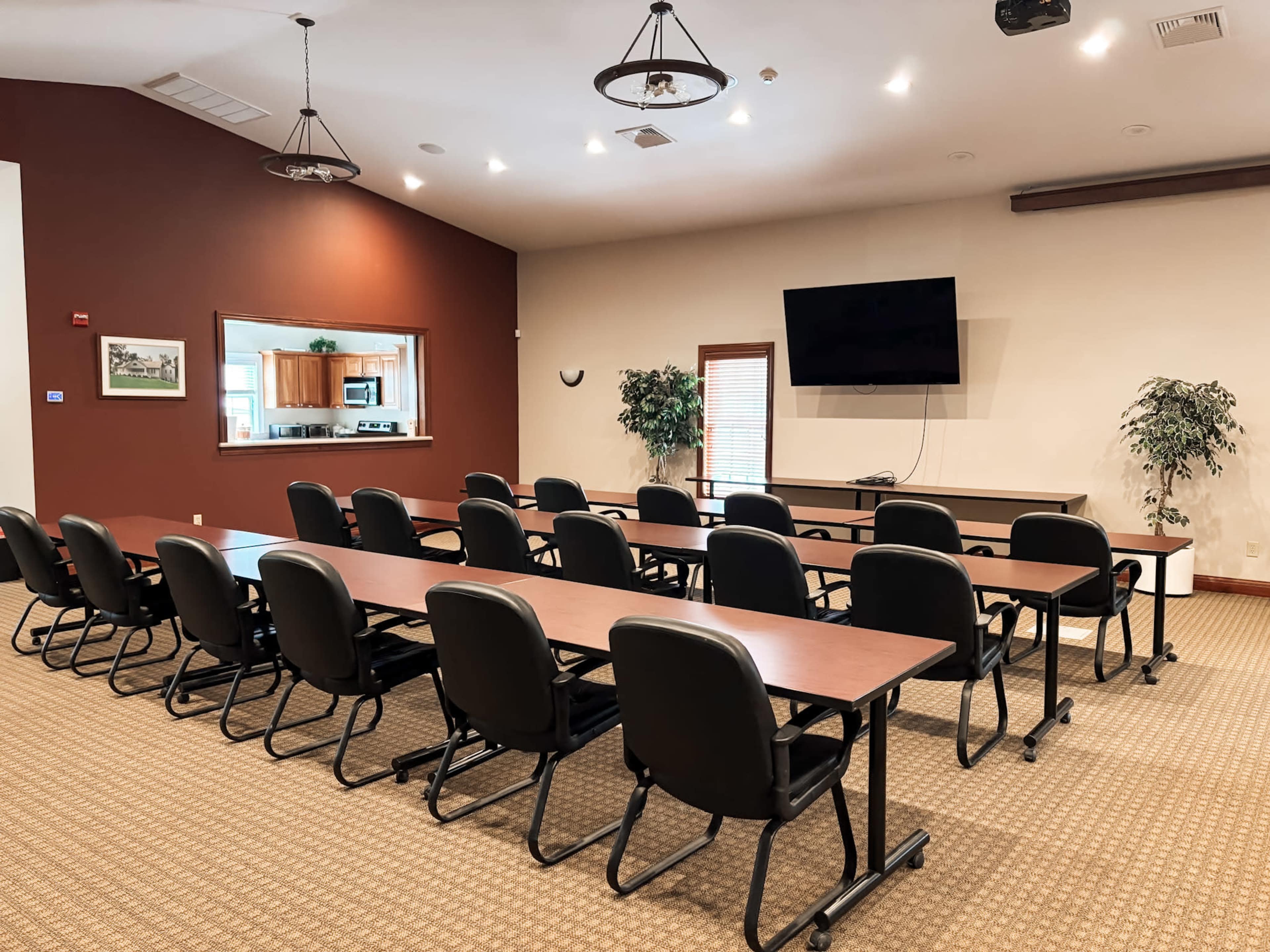 The image shows a spacious meeting room with several rows of chairs and tables arranged facing a wall-mounted television and a window opening to a kitchen area.