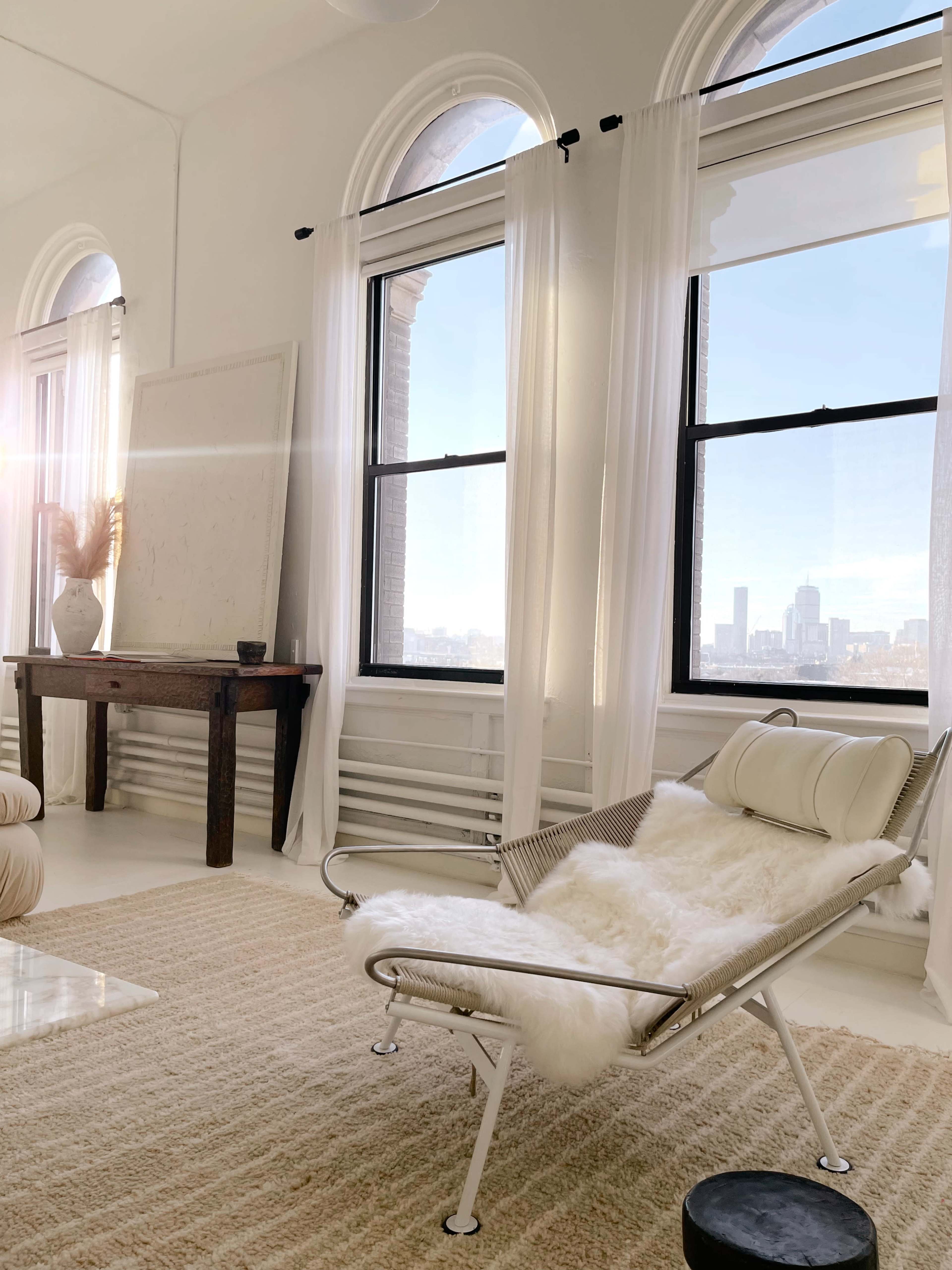 The scene features a minimalist living space with a sleek lounge chair covered in a fur throw, a wooden table, and large windows providing a view of the city skyline.