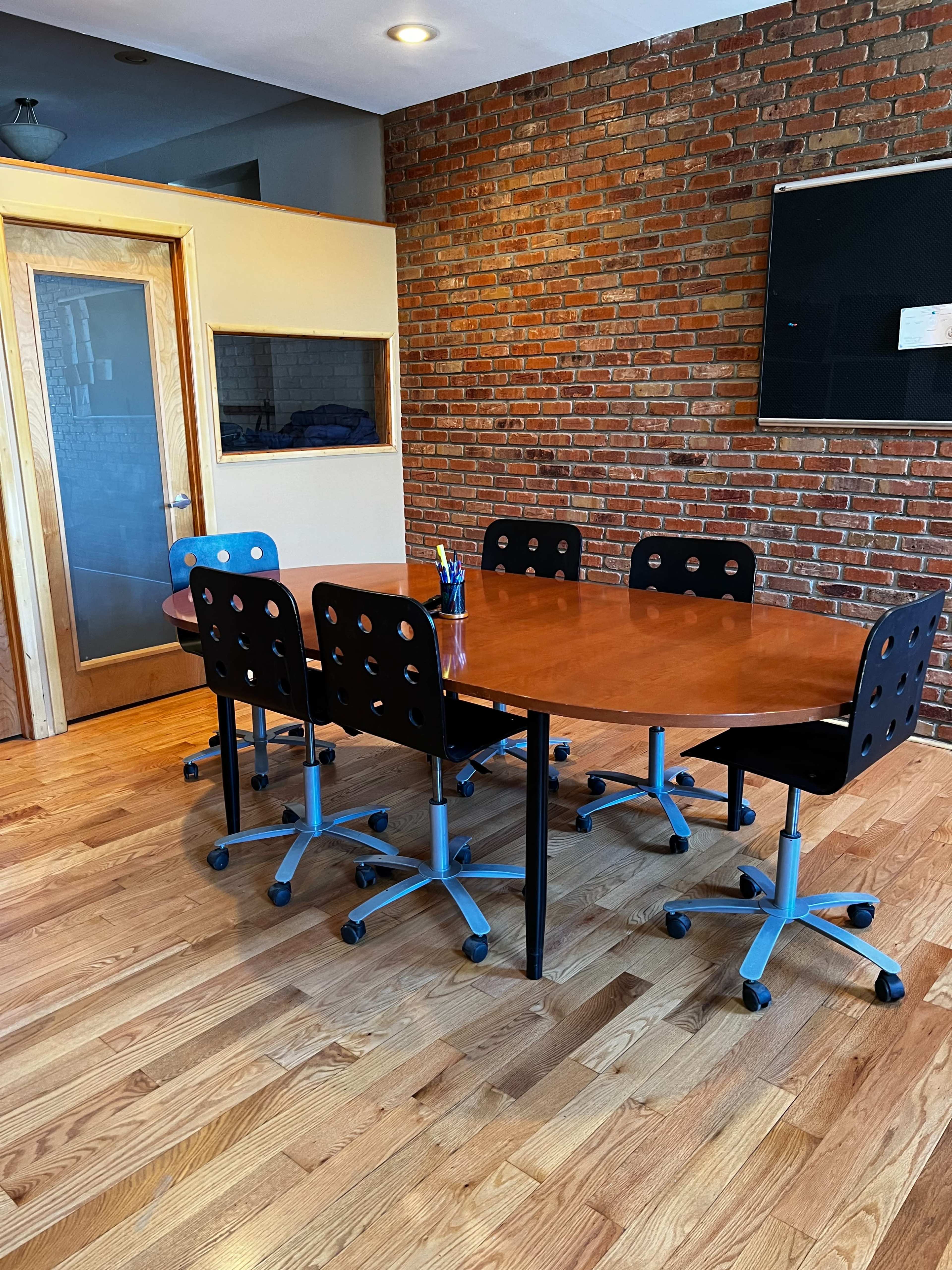 A conference room features a wooden table surrounded by six black office chairs on casters, with a brick wall in the background and a glass-panel door.