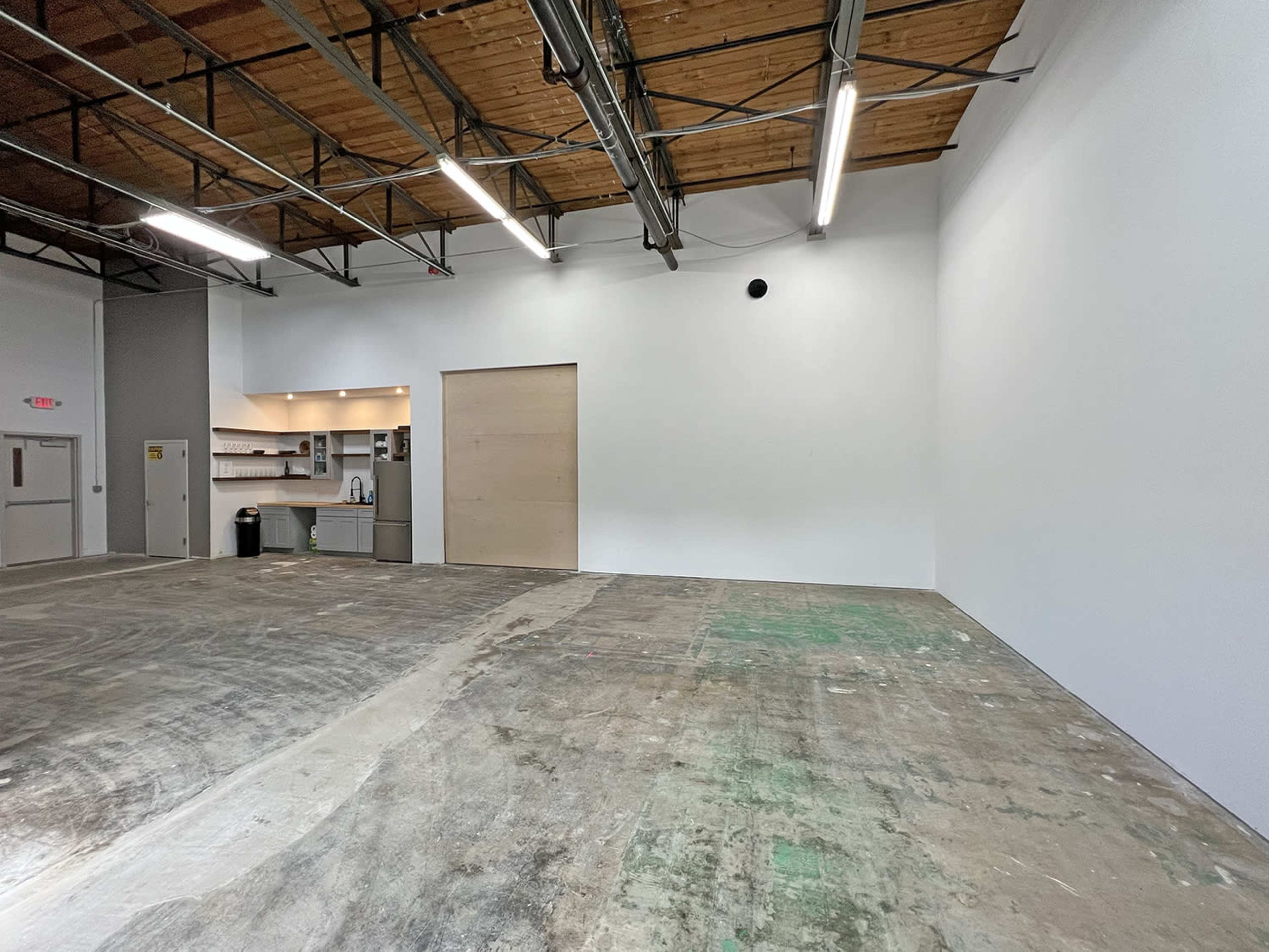An empty industrial space with a polished concrete floor, white walls, and a wooden door on one side leading to a kitchenette area.