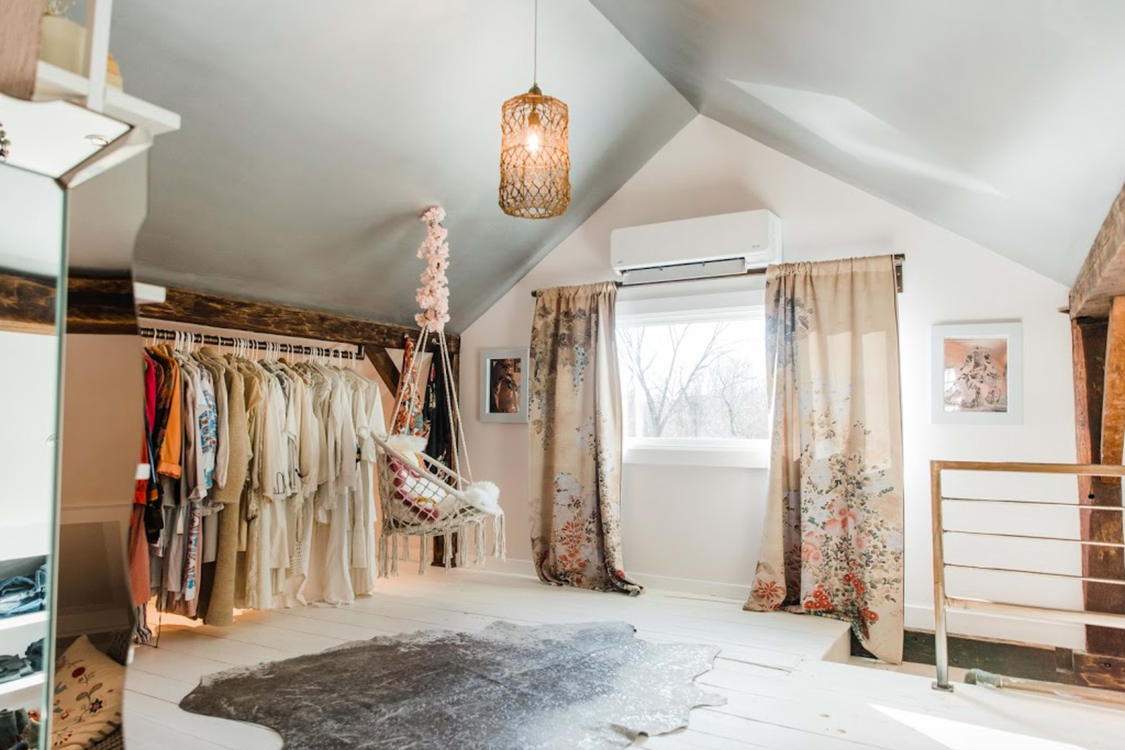 A well-lit attic bedroom with a hanging chair, a clothing rack filled with various garments, and large windows adorned with floral curtains.