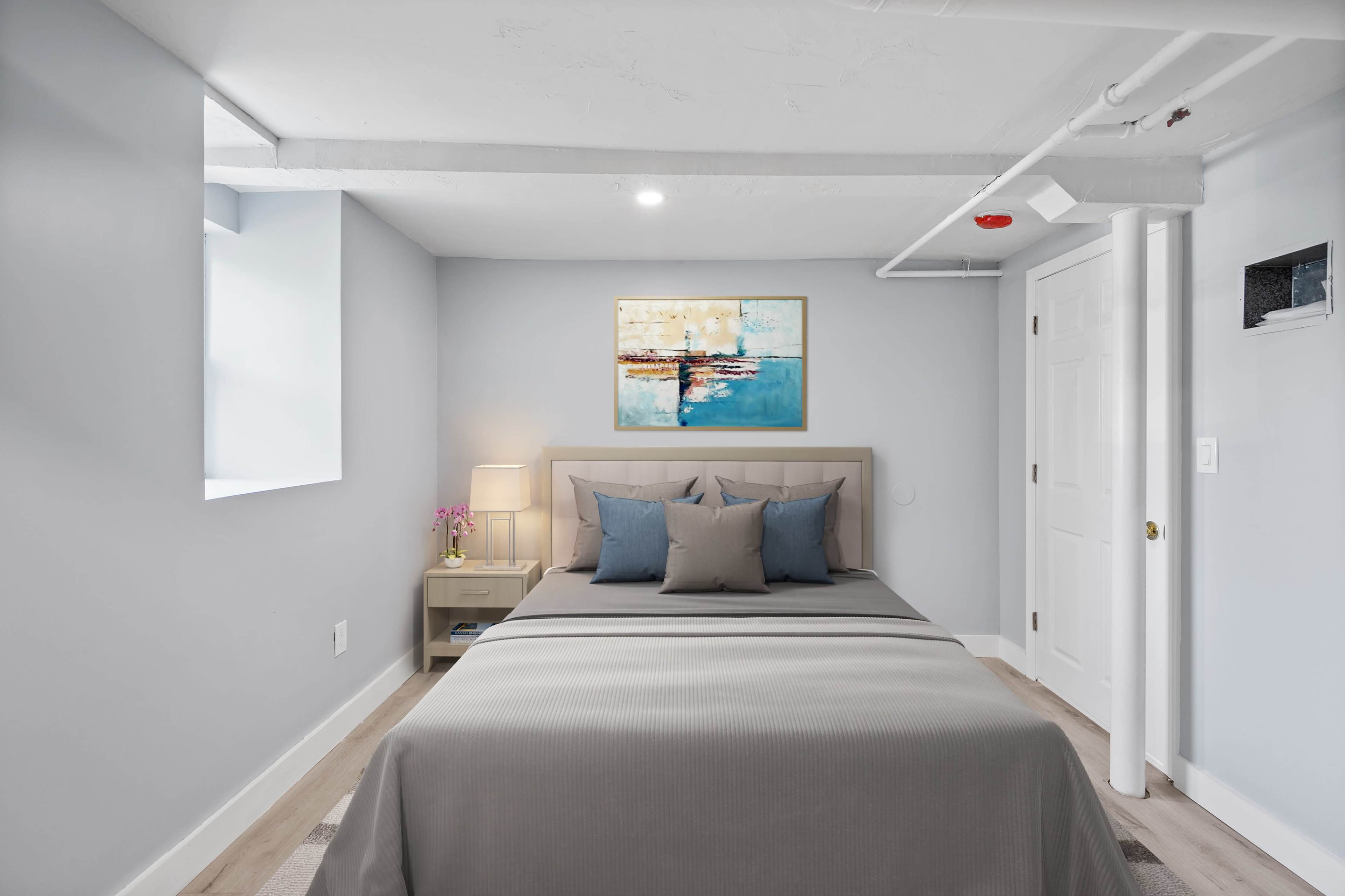 A well-lit bedroom featuring a bed with gray bedding, decorative pillows, and a nightstand with a lamp against a light blue wall.