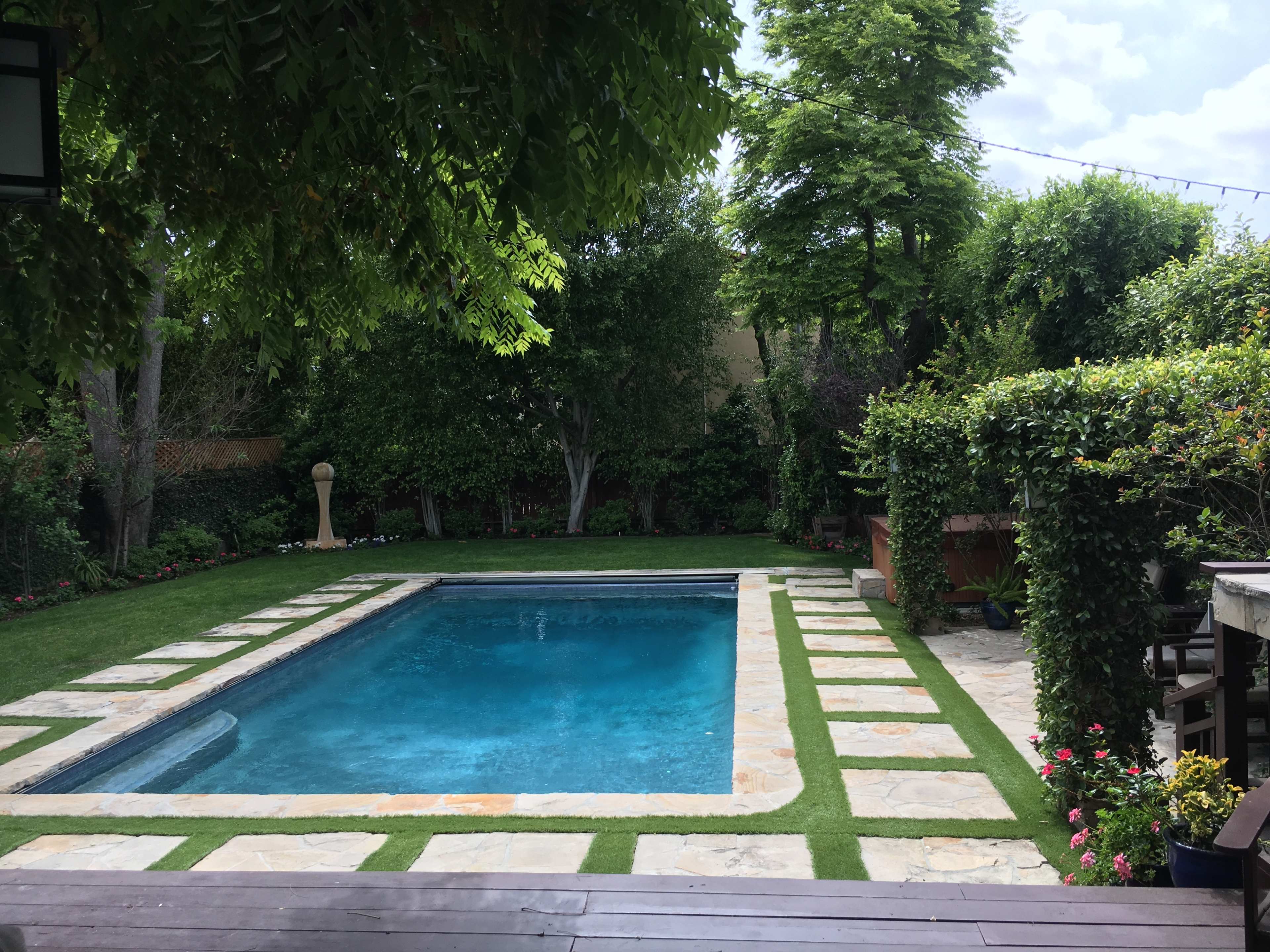 The image shows a neatly landscaped backyard featuring a rectangular swimming pool surrounded by stone pavers and lush greenery.