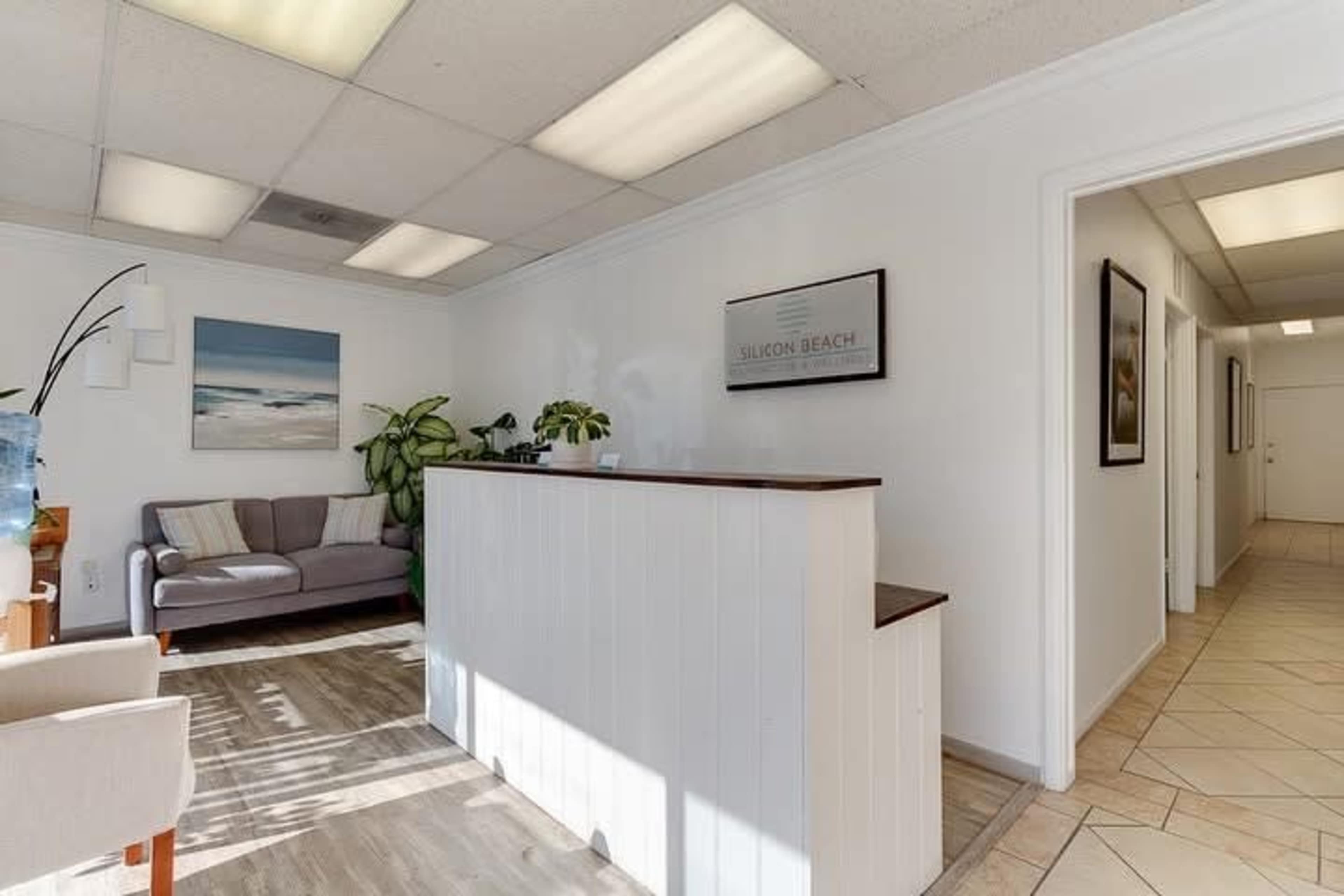 The image shows the interior of a reception area with a wooden front desk, a sofa, plants, and framed artwork on the walls.