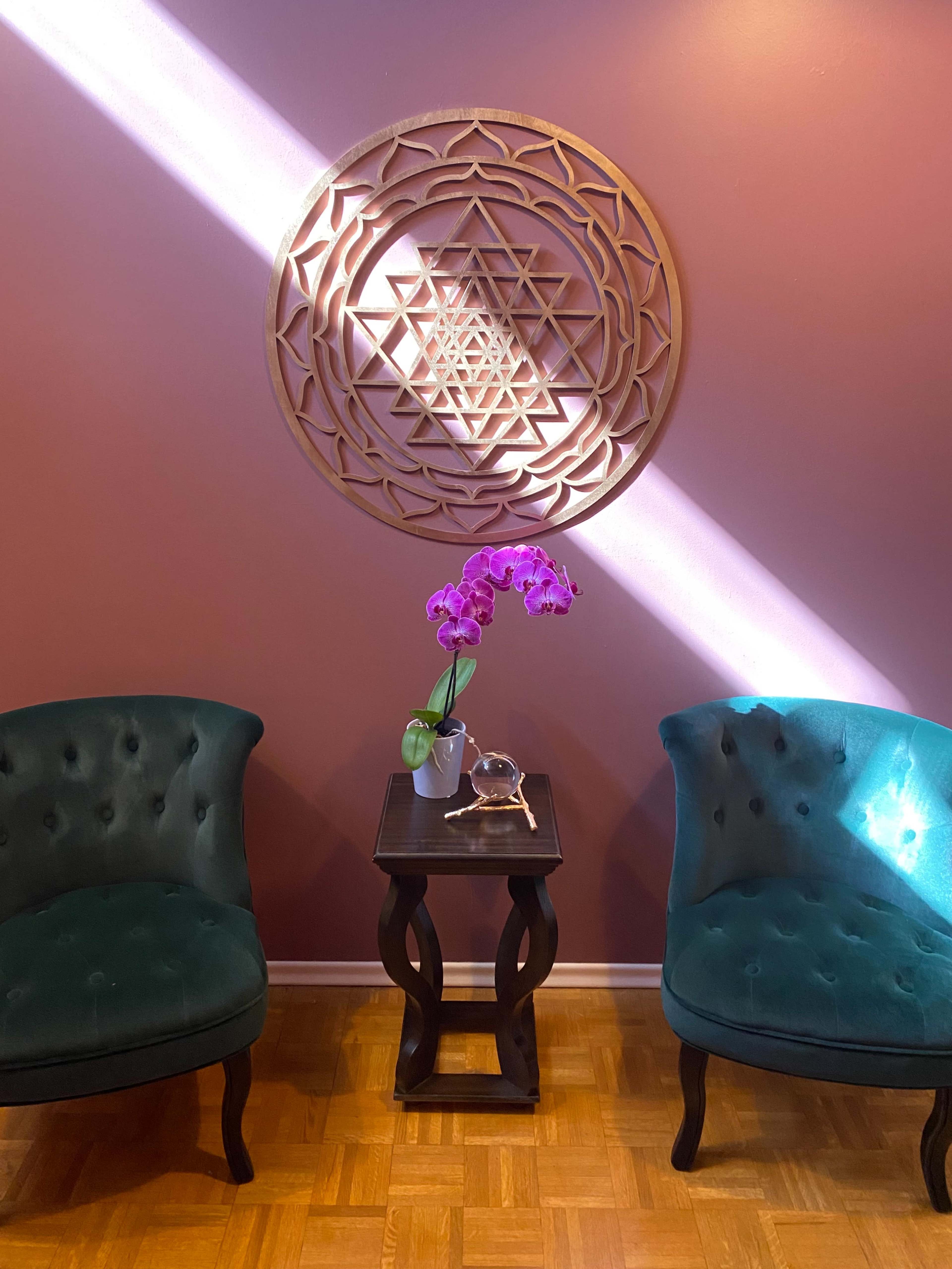 Two green, tufted chairs face each other across a dark wooden side table, which holds a small vase with an orchid and a decorative object, against a mauve wall featuring a circular mandala design.