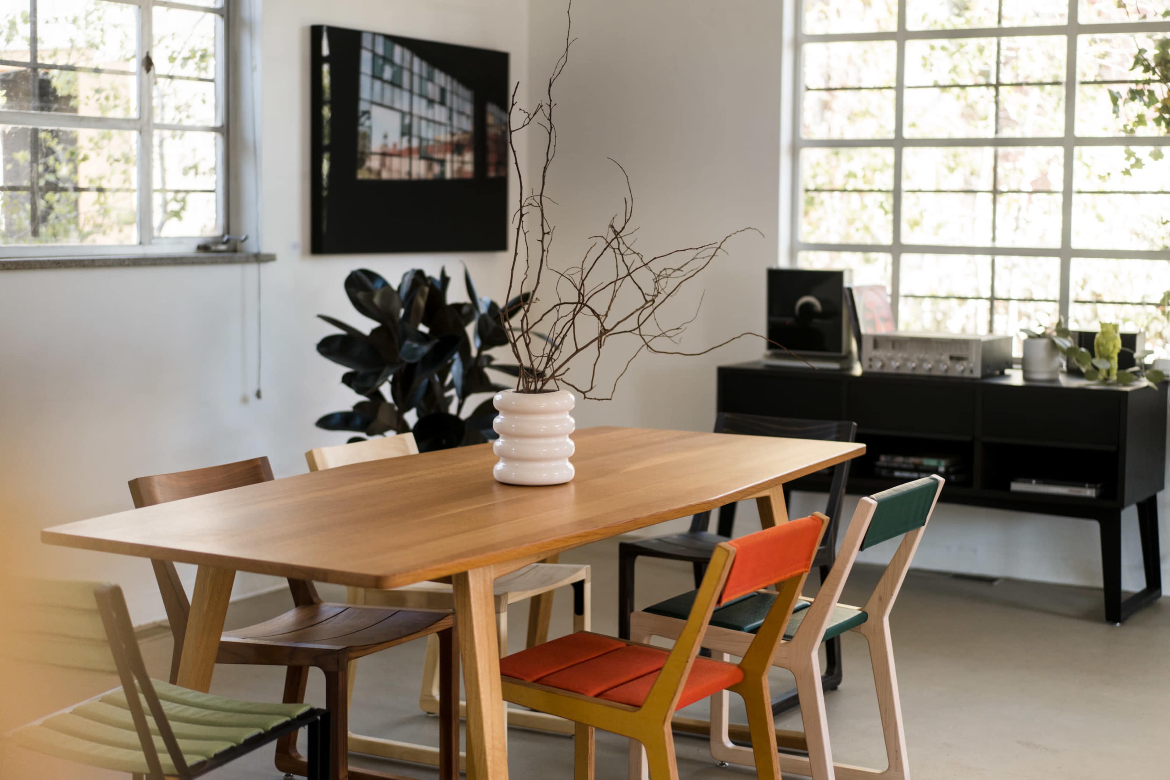 A wooden dining table with colorful chairs and a vase containing bare branches is set in a bright room featuring large windows and modern decor.