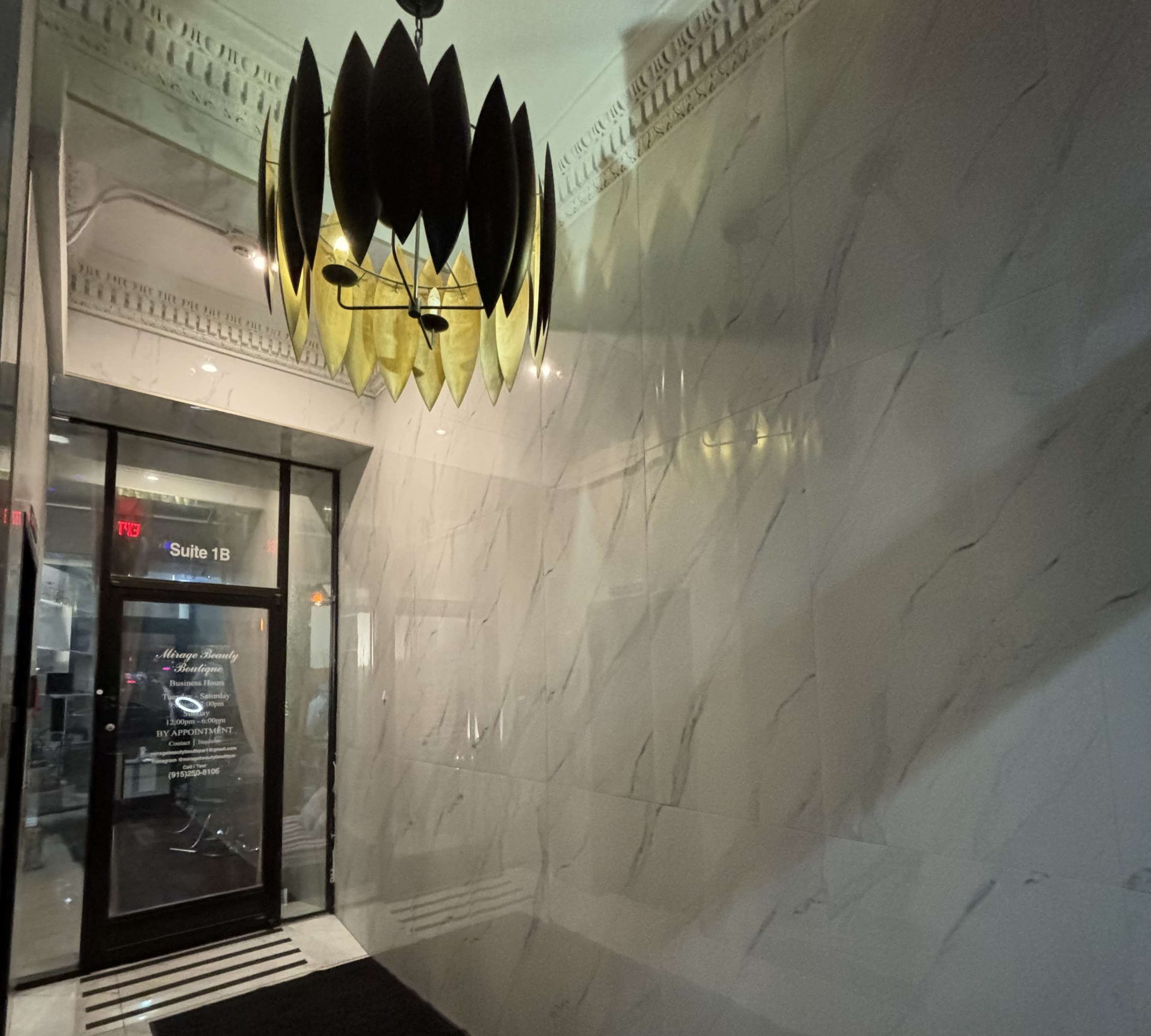 A modern entrance hallway features a glass door labeled "Suite 1B" and a large pendant light casting shadows on the marble walls.