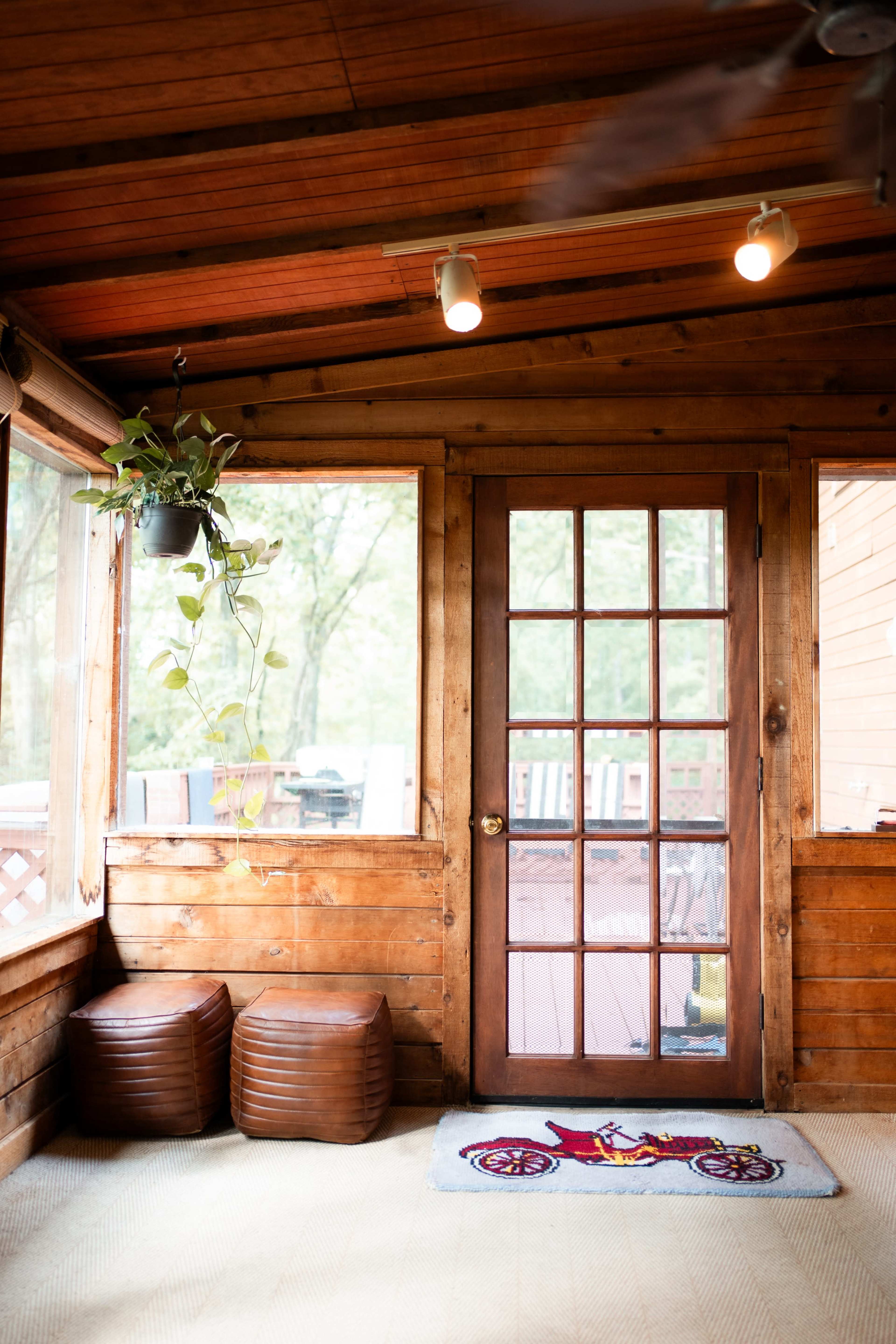 The image shows a wooden sunroom with a glass door, two brown ottomans, and a hanging plant.