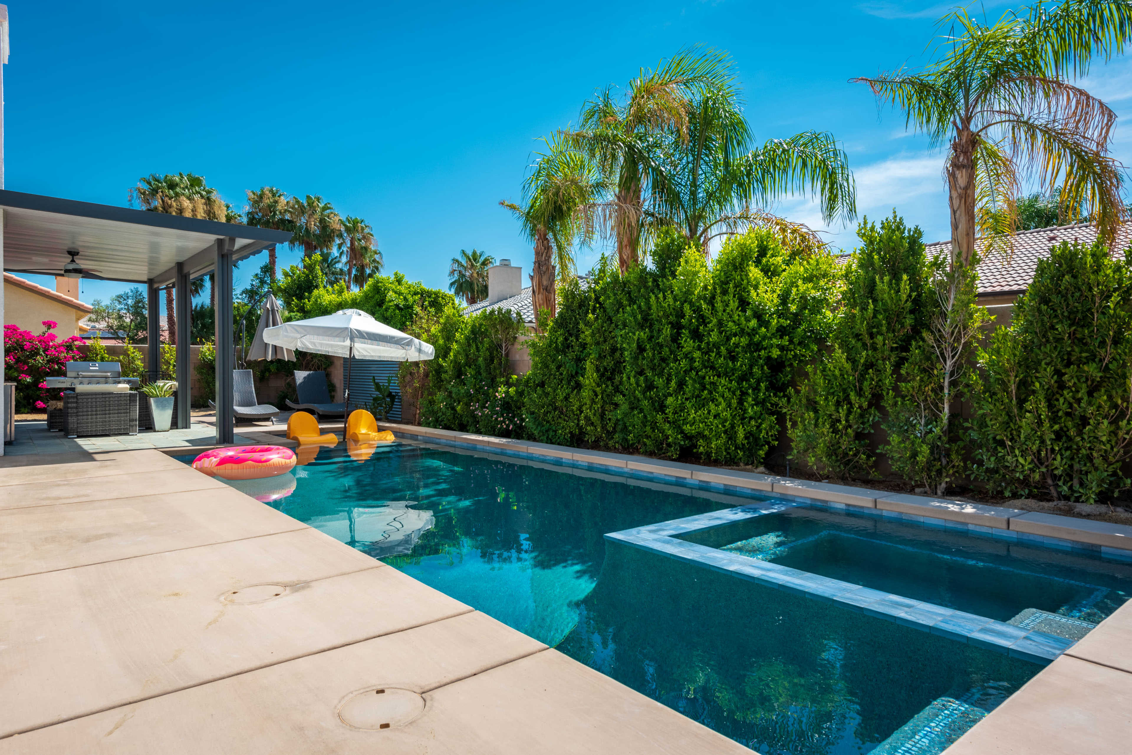 The image shows a backyard pool surrounded by palm trees, shrubs, and outdoor furniture, with inflatable floats on the water.