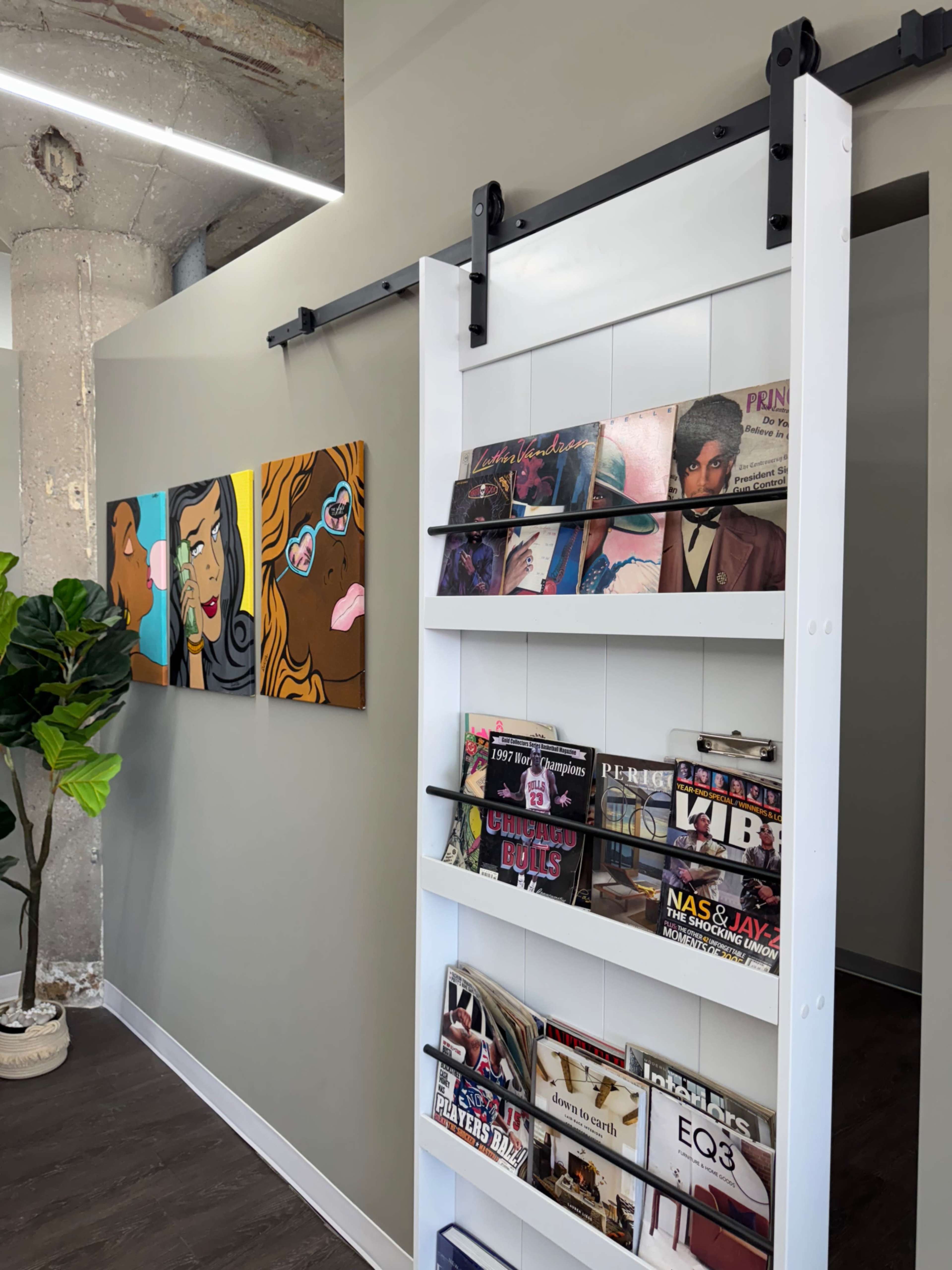 The image shows a modern interior with a sliding bookcase displaying magazines and two colorful art pieces on the wall.