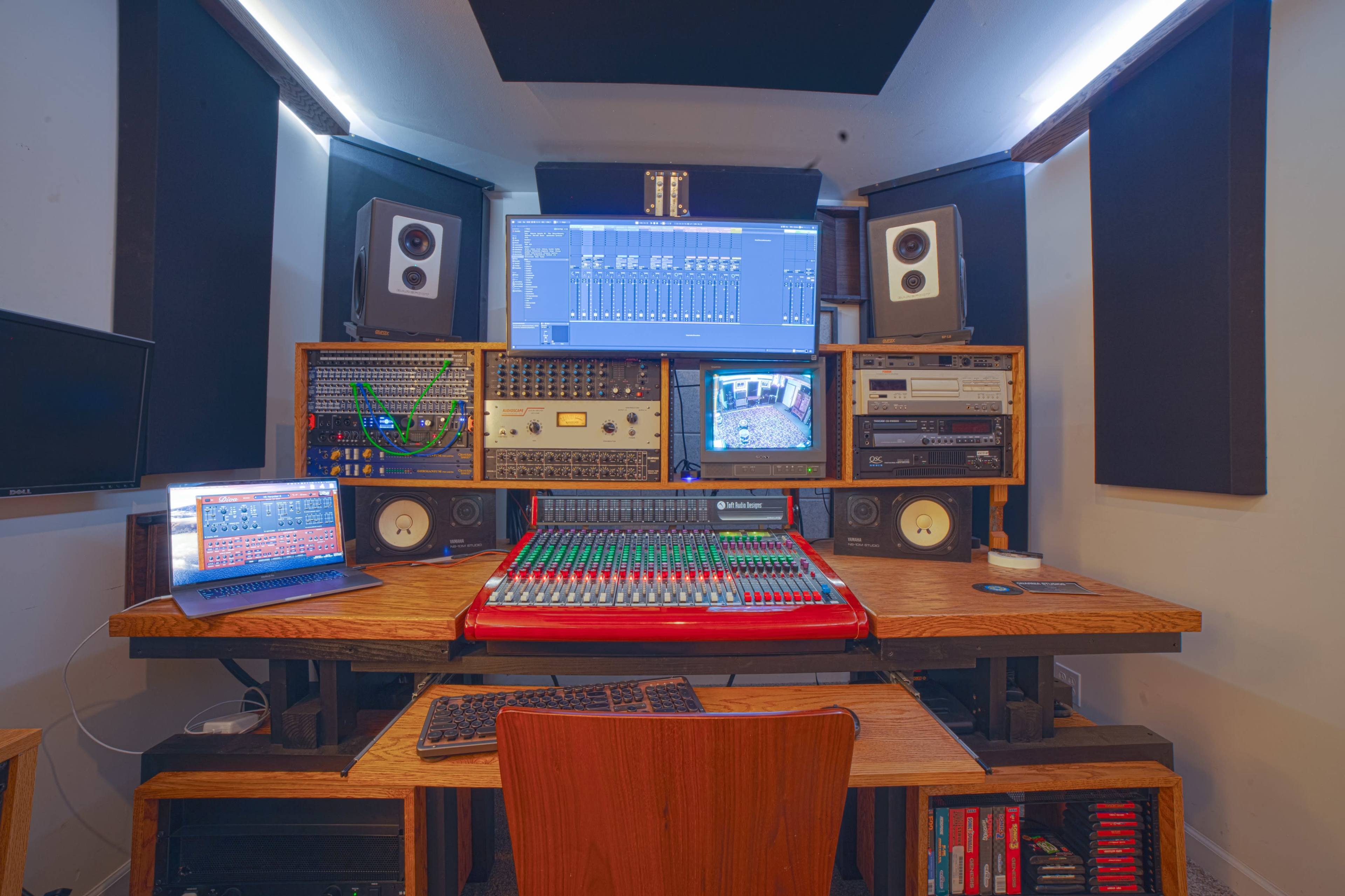 The image shows a music production studio featuring a central mixing console, multiple monitors, speakers, and various audio equipment arranged around a wooden desk.