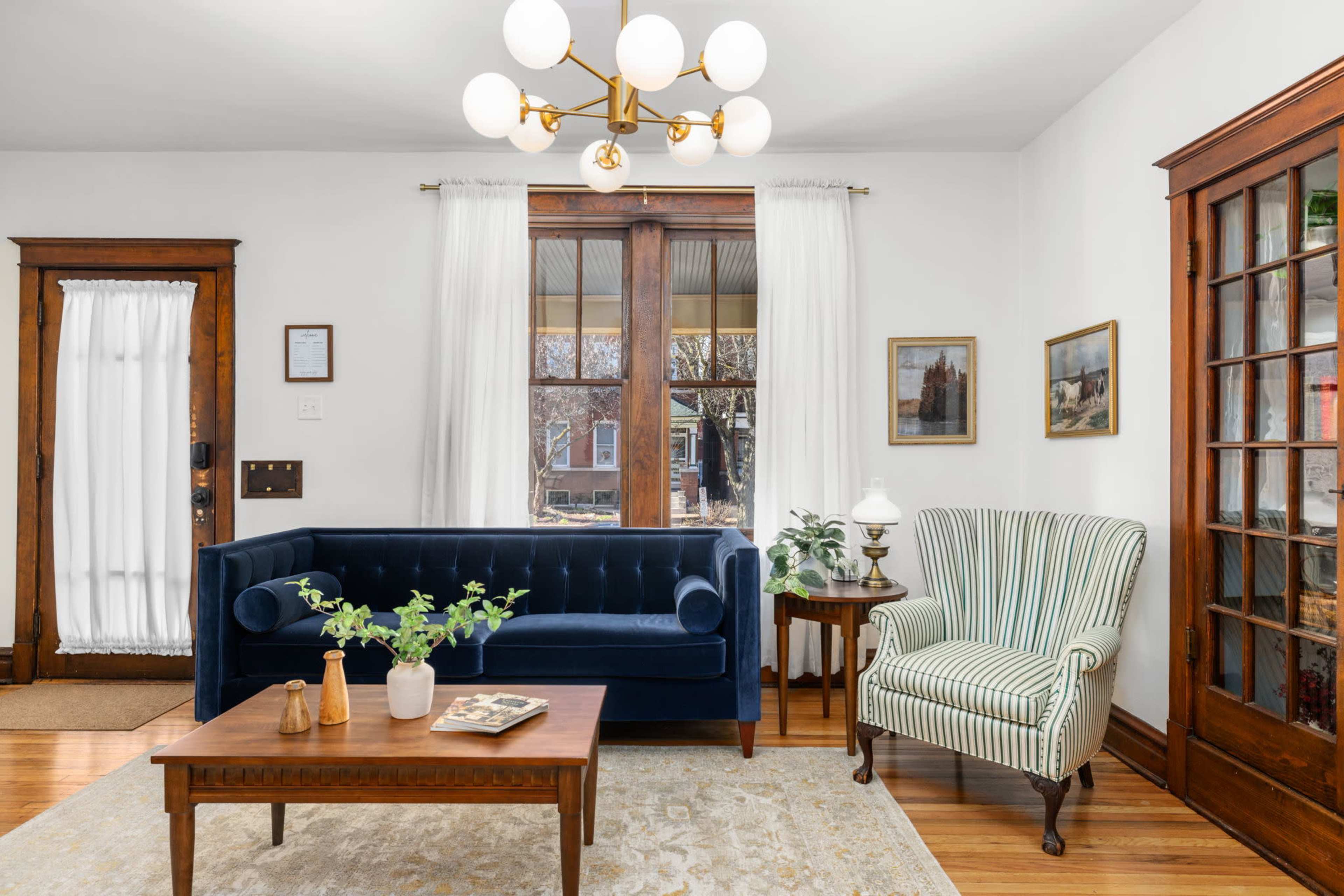 The image depicts a cozy living room featuring a blue velvet sofa, a striped armchair, a wooden coffee table, and large windows adorned with sheer curtains.