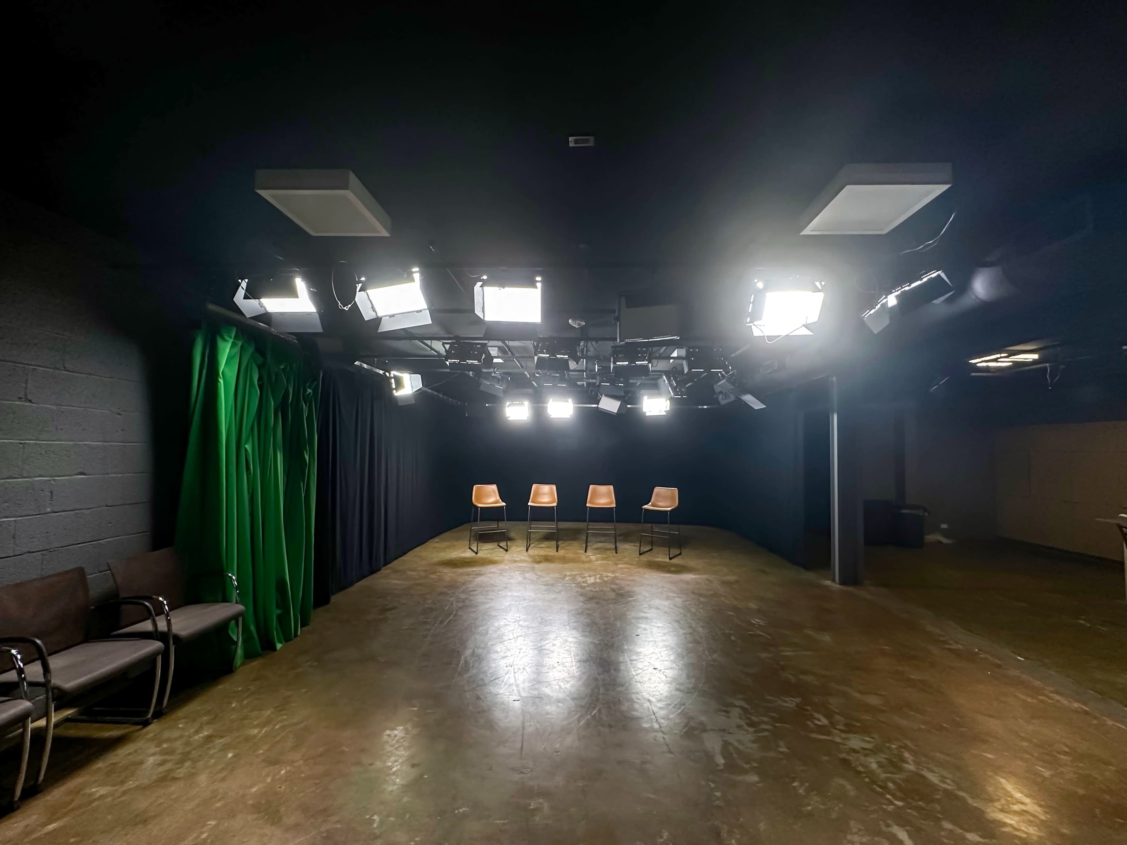 A dimly lit studio features four empty chairs positioned in a semicircle, surrounded by bright overhead lights and black curtains.