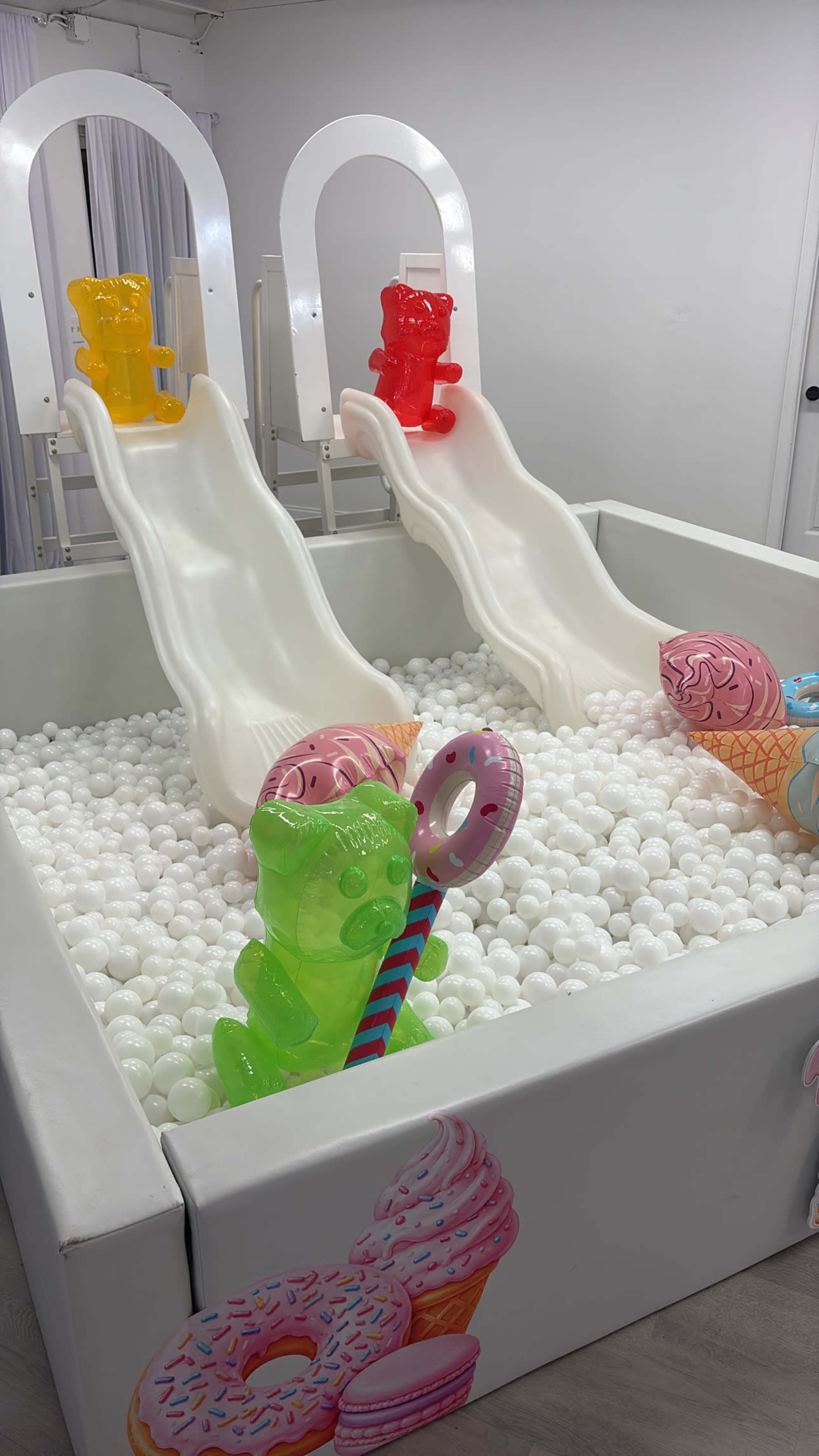 The image shows an indoor play area featuring two white slides surrounded by a pit filled with white plastic balls and colorful inflatable toys.
