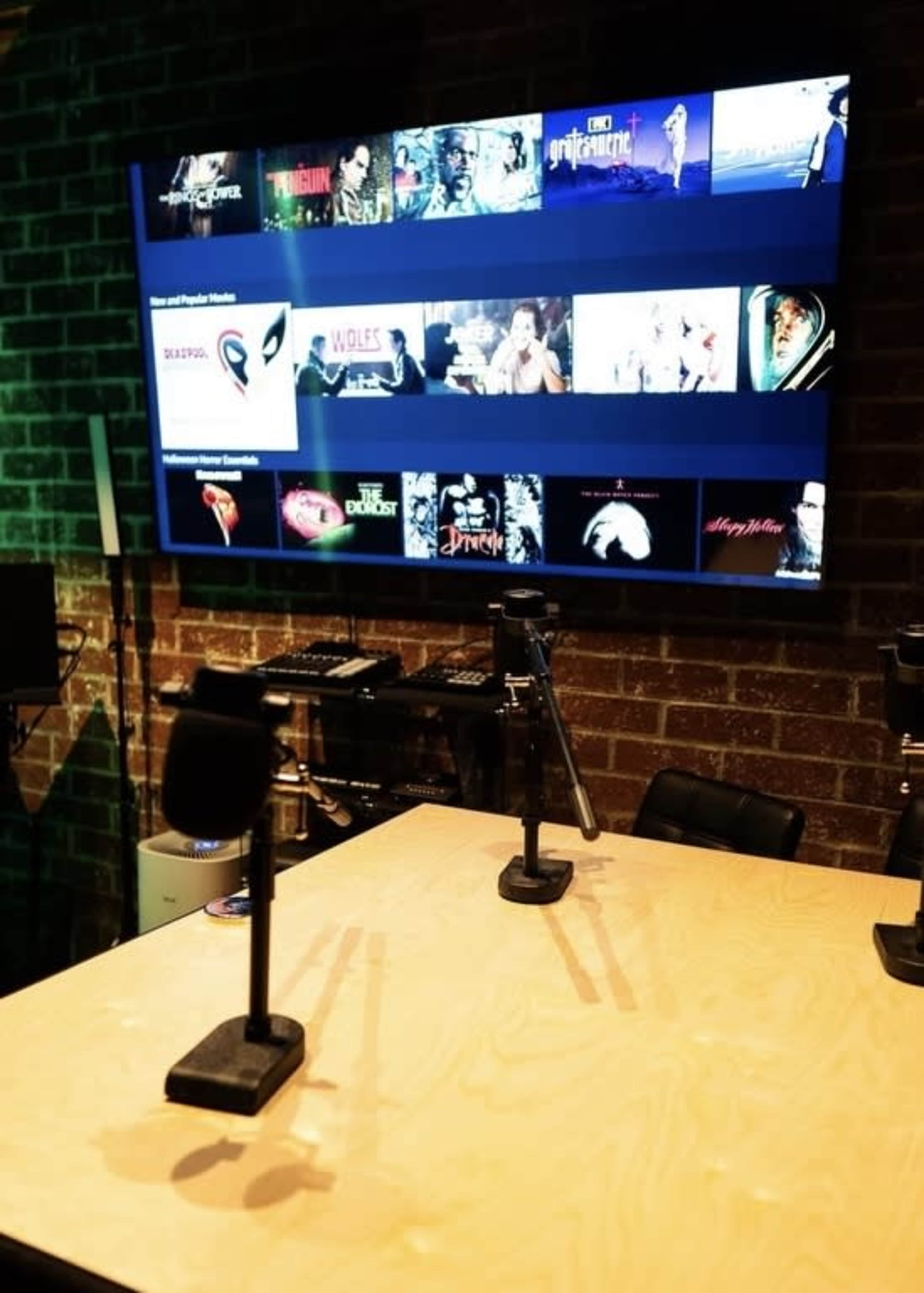 The image shows a recording studio setup with a wooden table and two microphone stands, alongside a large screen displaying various movie titles.