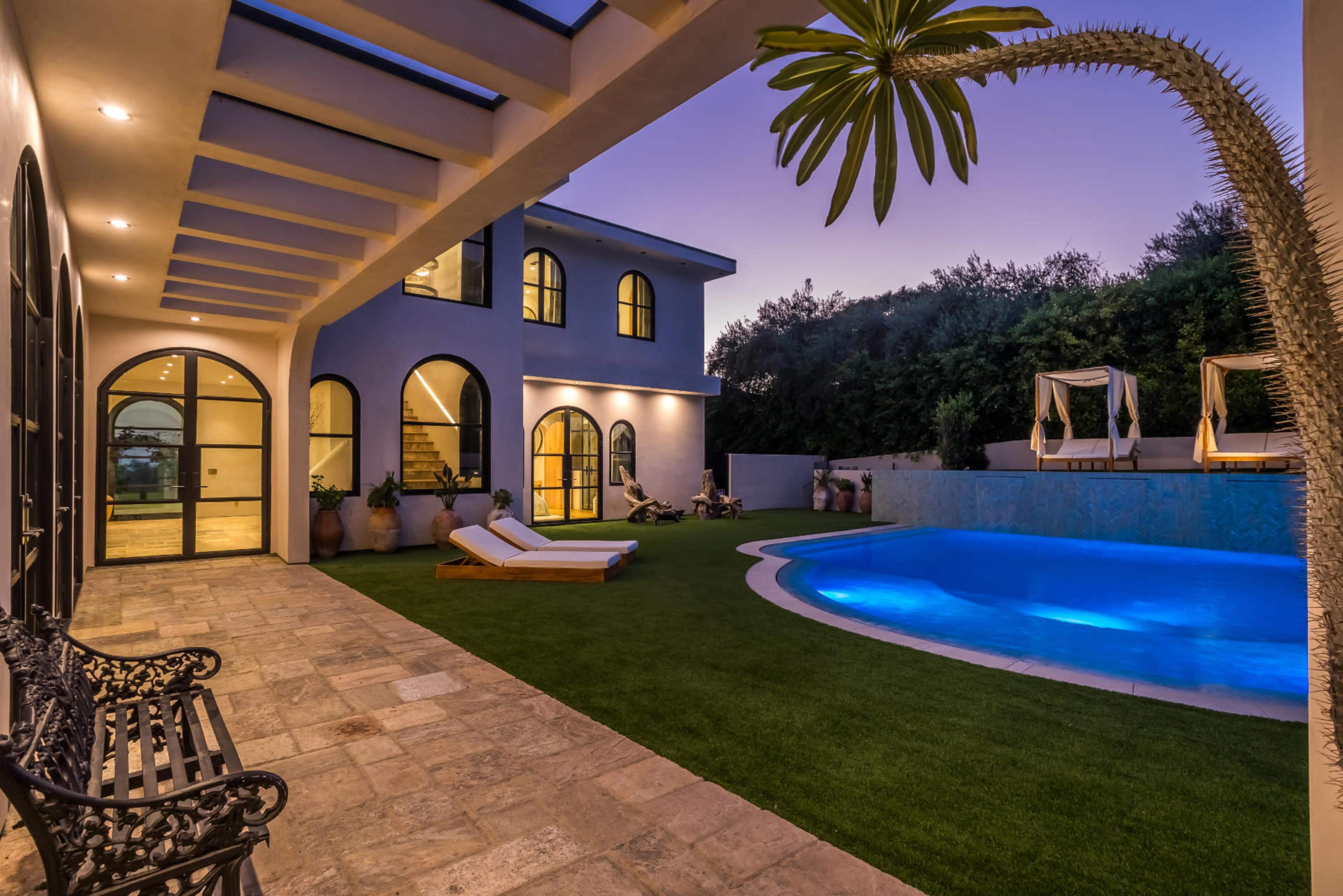 The image shows a modern villa with a swimming pool, lounge chairs, and an outdoor seating area under a covered patio at dusk.