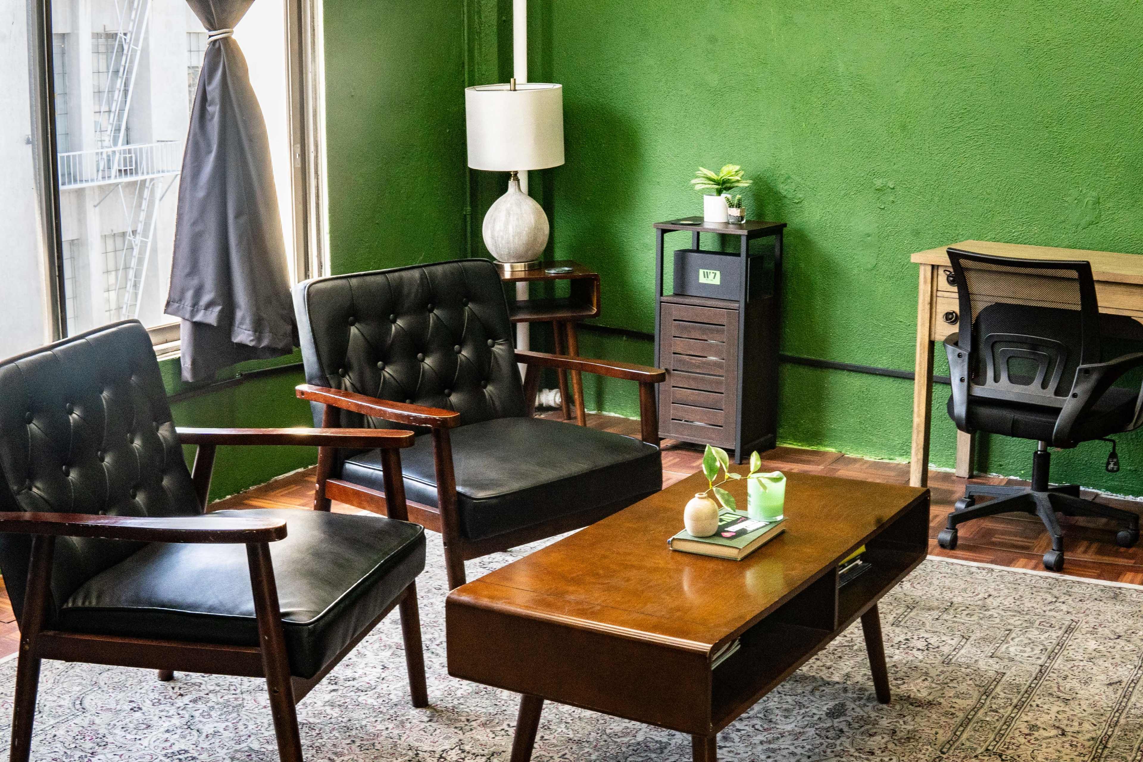 The image shows a cozy sitting area with two black armchairs, a wooden coffee table, a small plant, a lamp, and a desk against a green wall.