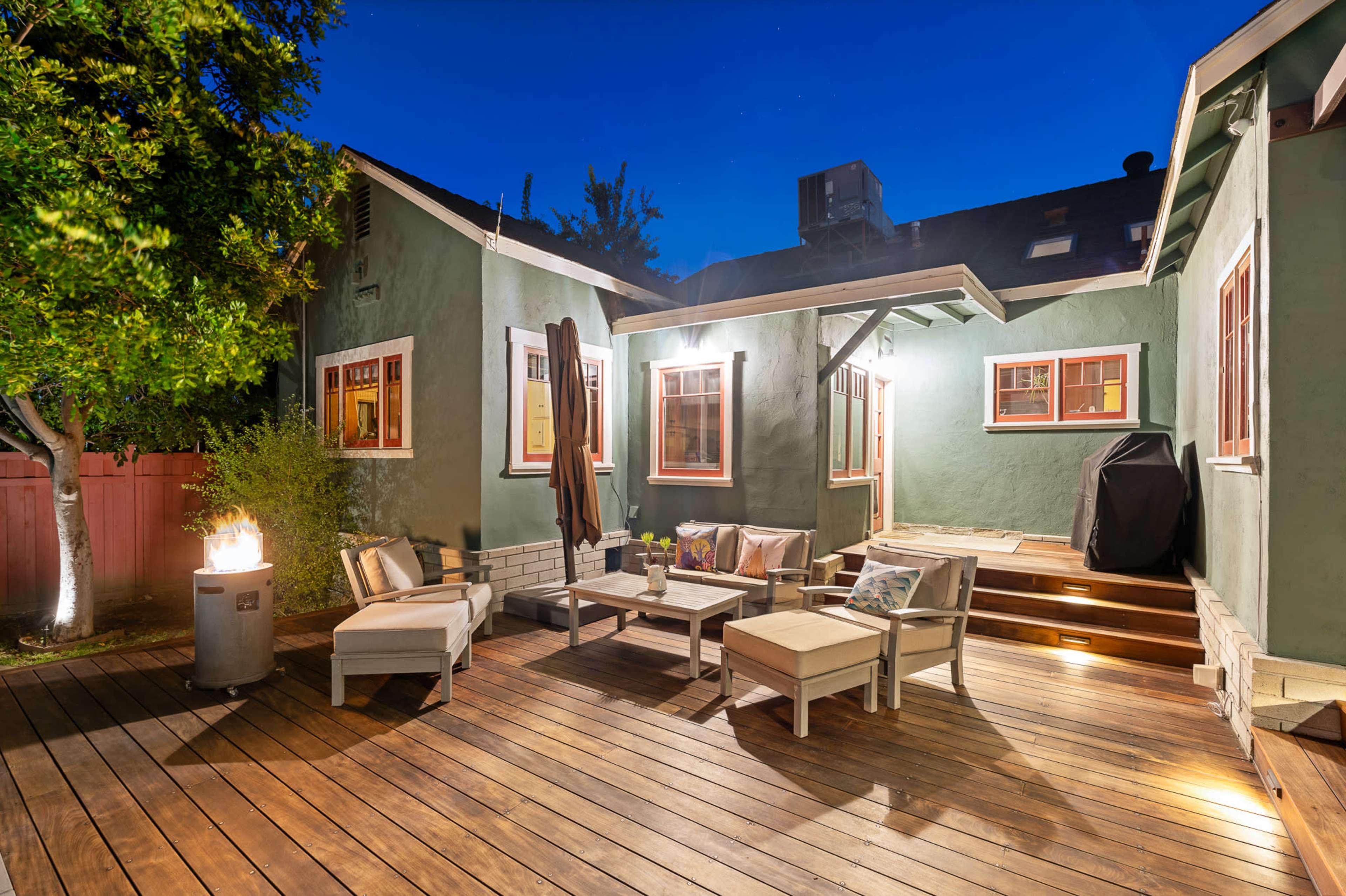 A cozy outdoor seating area with wooden furniture and a grill is illuminated by soft lighting in the evening.