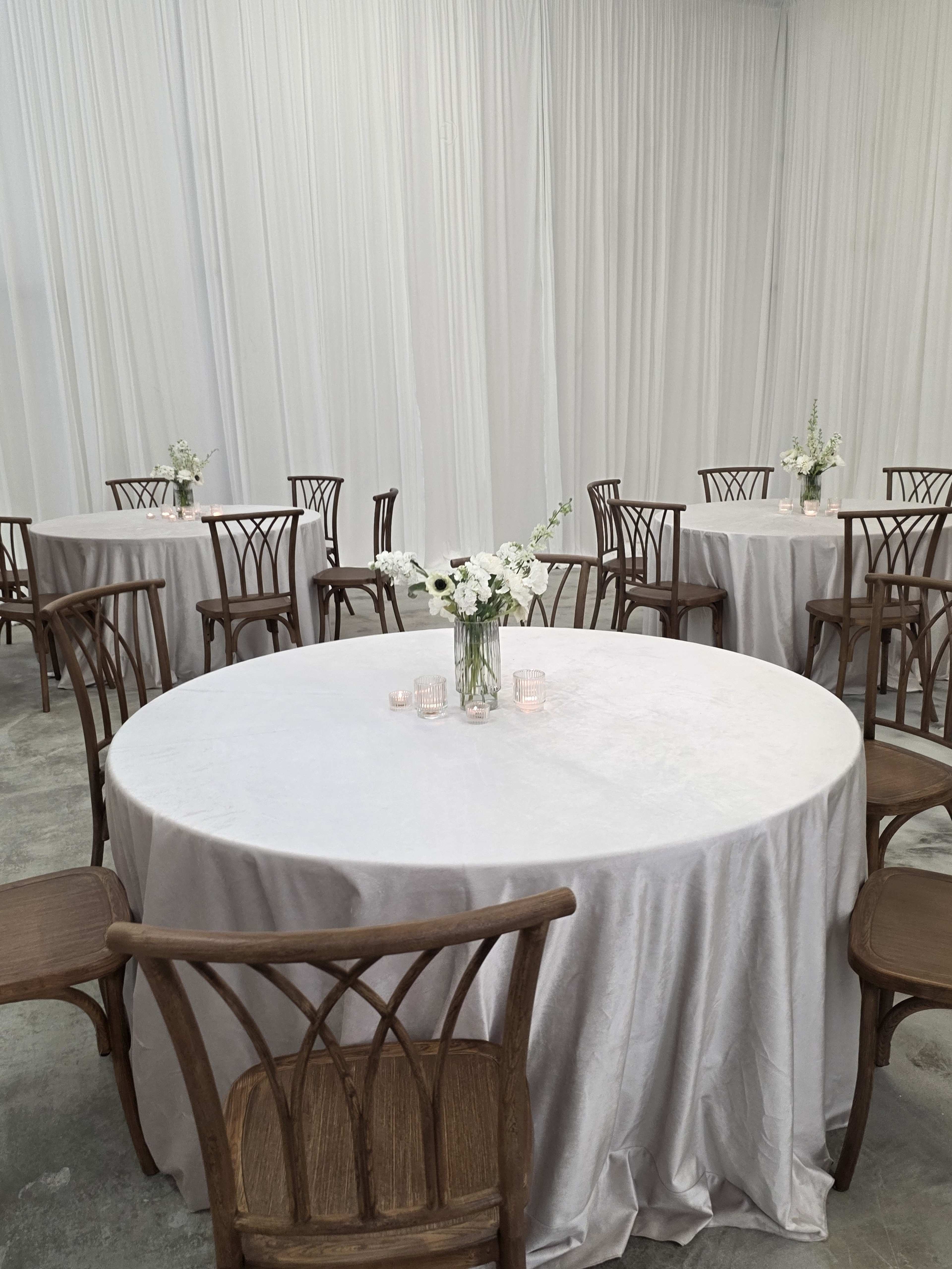 A room with several round tables covered in gray tablecloths, each adorned with a small vase of white flowers and surrounded by wooden chairs.