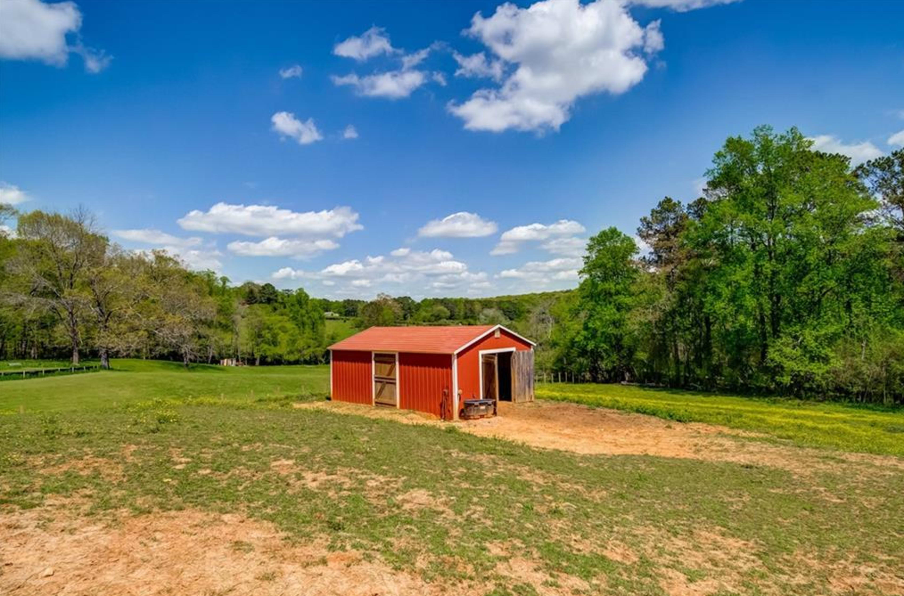 A red barn is situated in a grassy field surrounded by trees under a blue sky with scattered clouds.