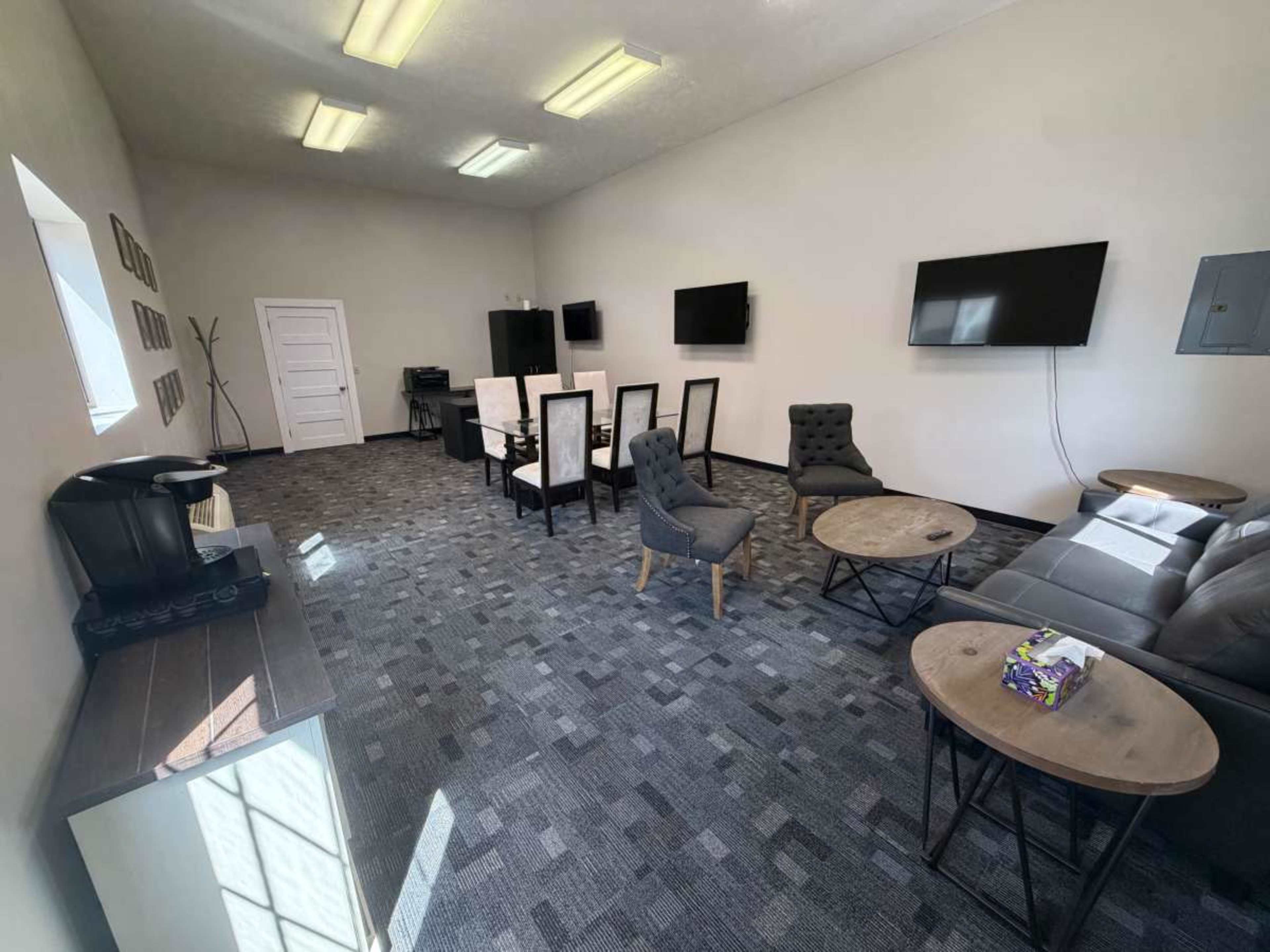 The image shows a spacious, modern waiting room with several chairs, tables, and multiple wall-mounted televisions.