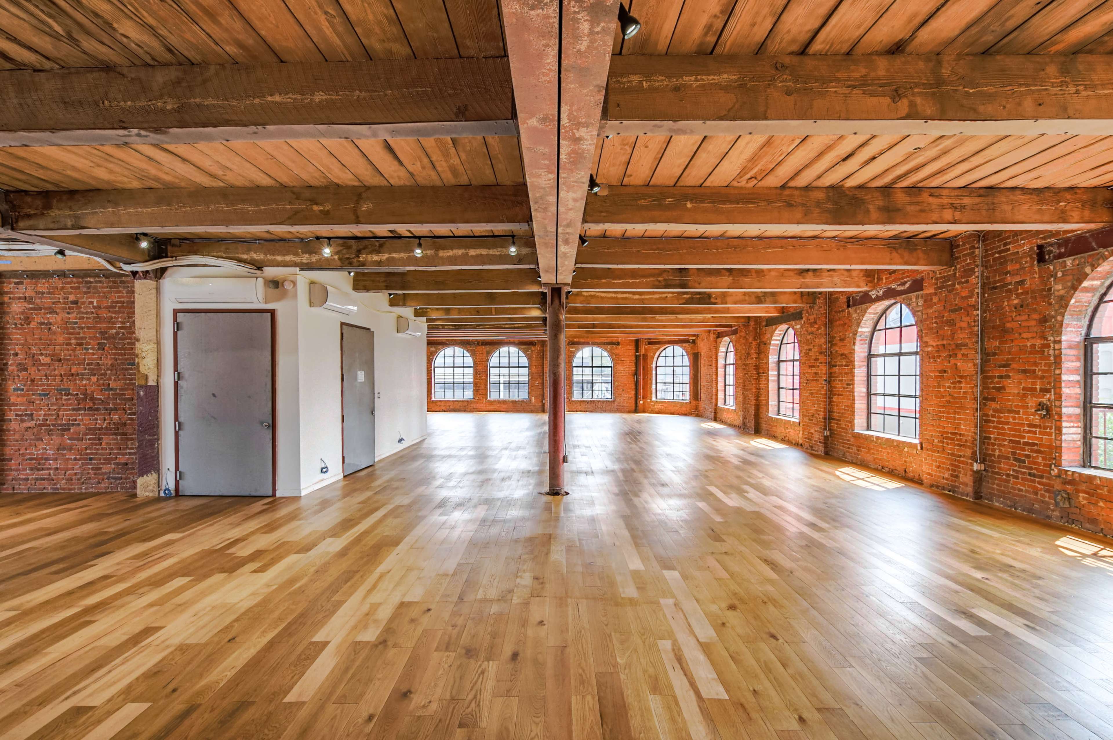 The image shows a spacious, well-lit interior featuring wooden beams, a polished wooden floor, and large arched windows along the brick walls.
