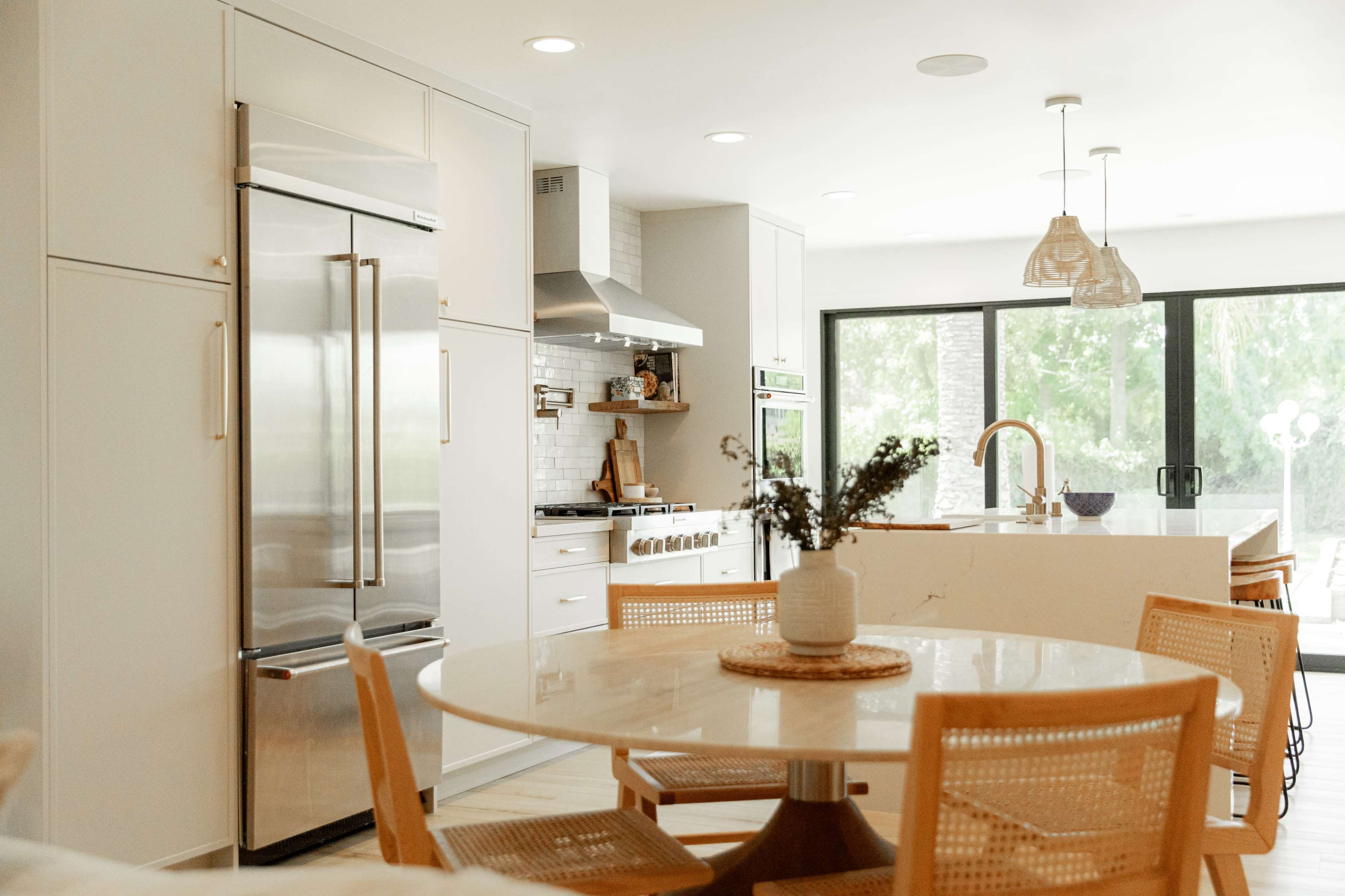 A modern kitchen features stainless steel appliances, a large island with seating, and a dining table surrounded by woven chairs.