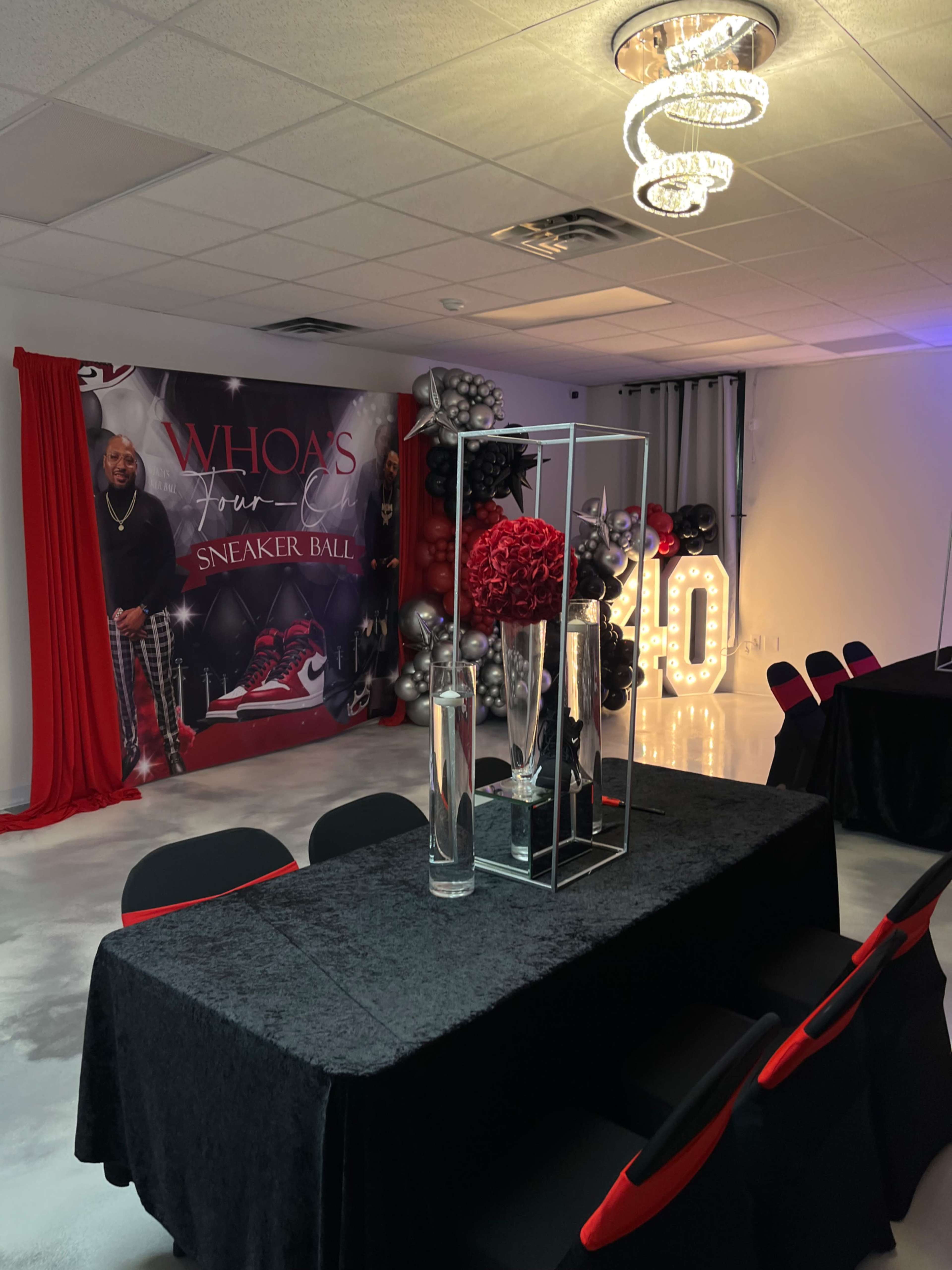 A decorated indoor venue features a black and red color scheme with a table set for an event, surrounded by balloons and a backdrop for a sneaker-themed celebration.