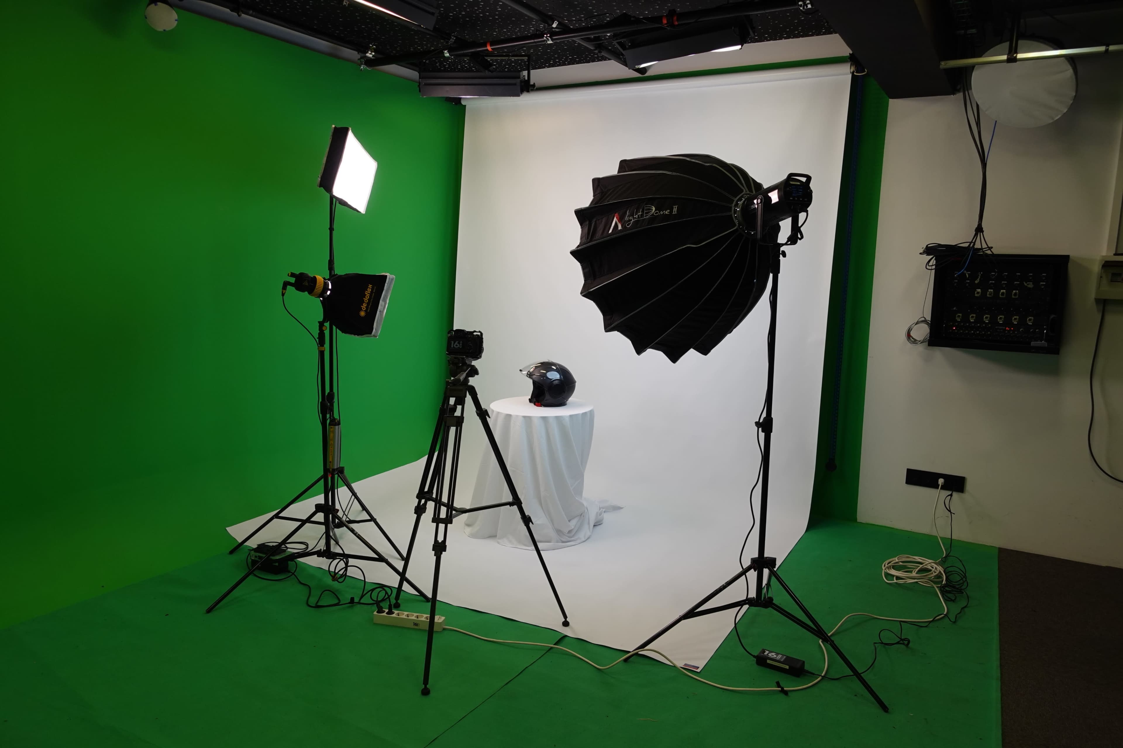 A photography studio setup with a green backdrop, a softbox light, a camera on a tripod, and a helmet placed on a pedestal.