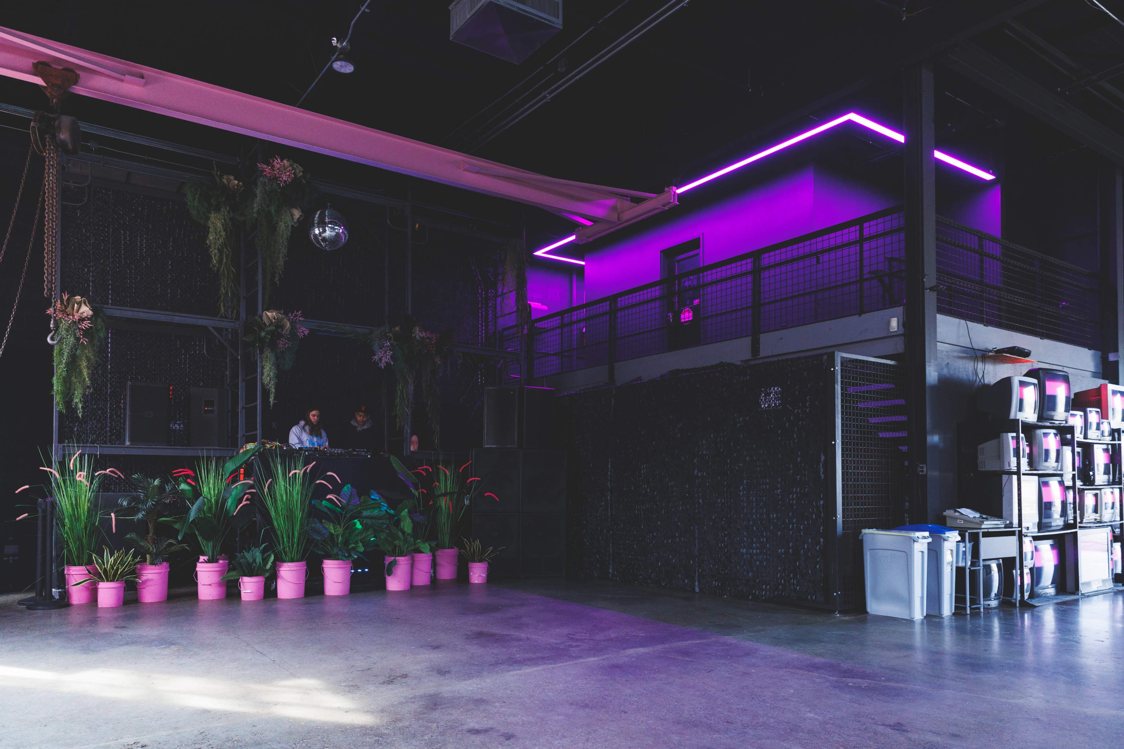 A modern indoor space with a dark interior, highlighted by purple neon lights, potted plants in pink containers, and a staircase leading to an upper level.
