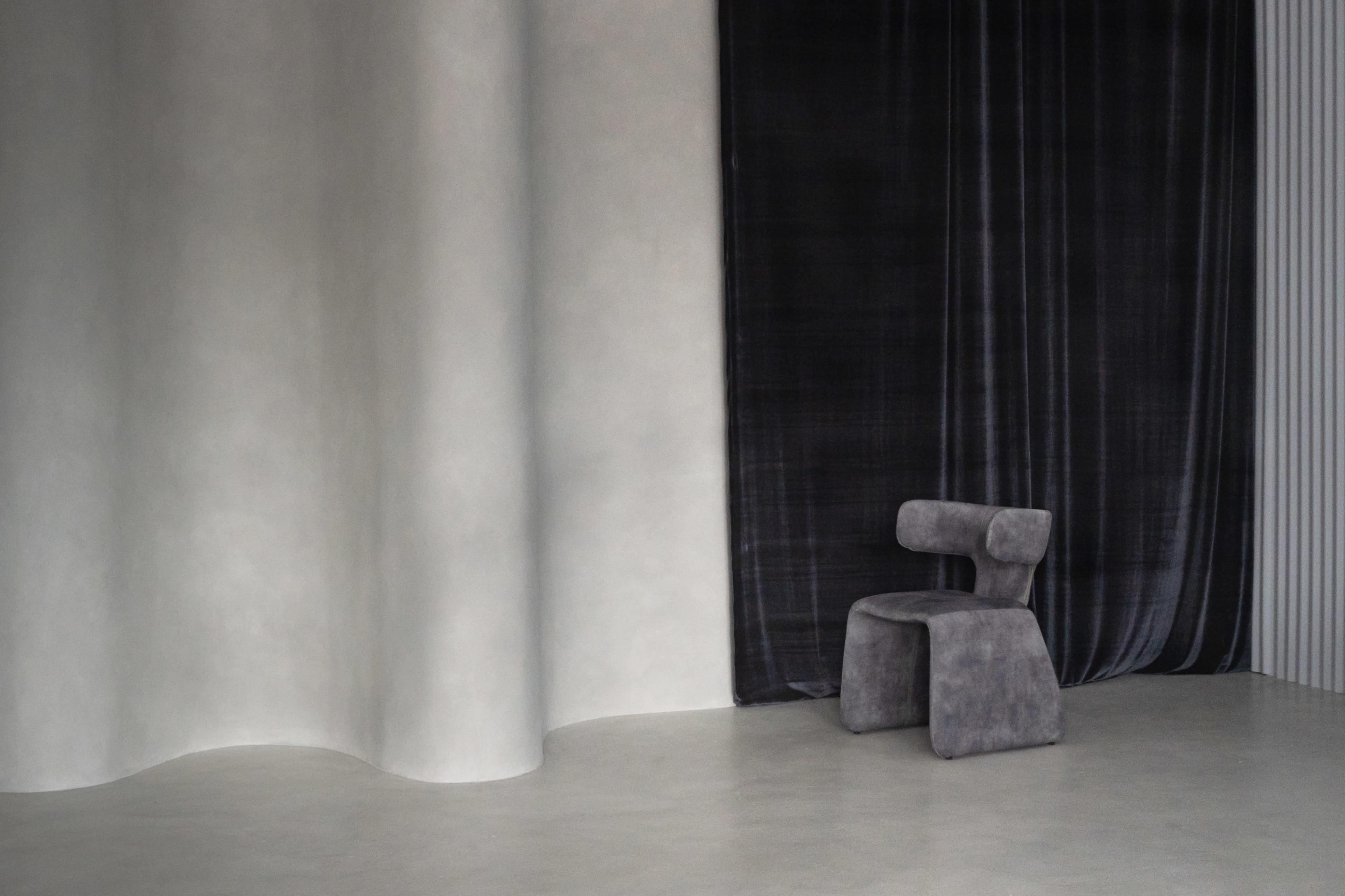 A modern, minimalist interior features a gray chair against a backdrop of smooth, curved walls and dark fabric draping.