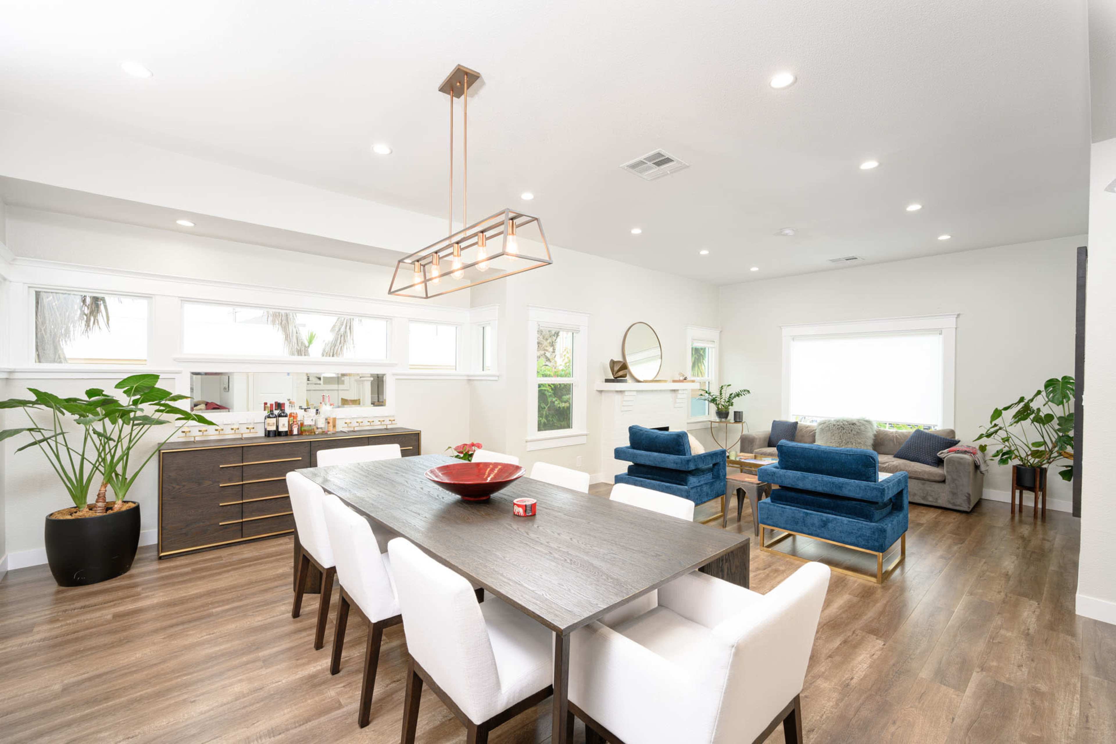A modern open-concept living and dining area features a wooden dining table with white chairs, a sleek sideboard, and a spacious seating arrangement with blue and gray sofas.