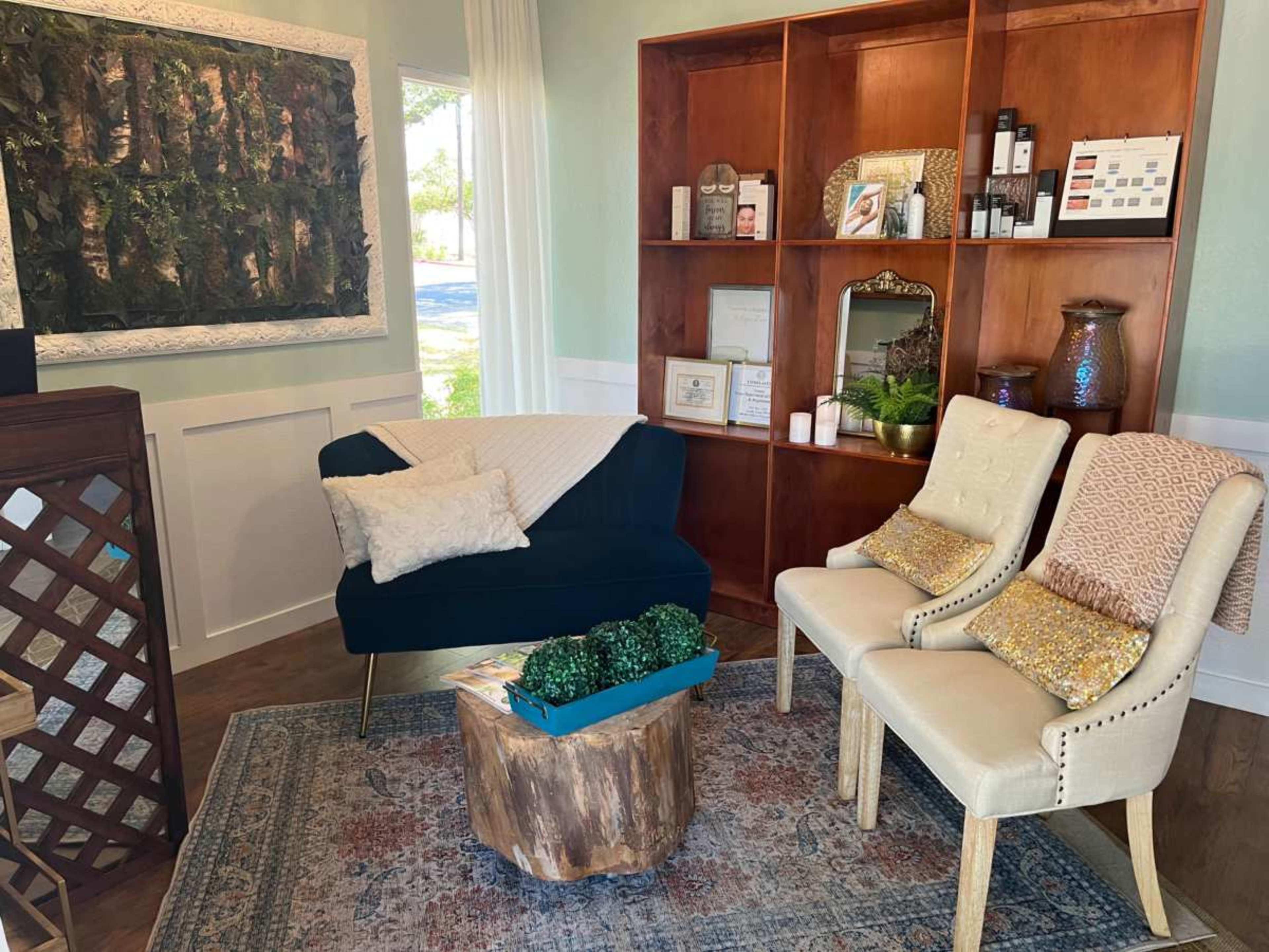 The image depicts a cozy interior scene featuring a dark blue couch, two cream-colored chairs, a wooden coffee table, and a shelf filled with decorative items and skincare products.
