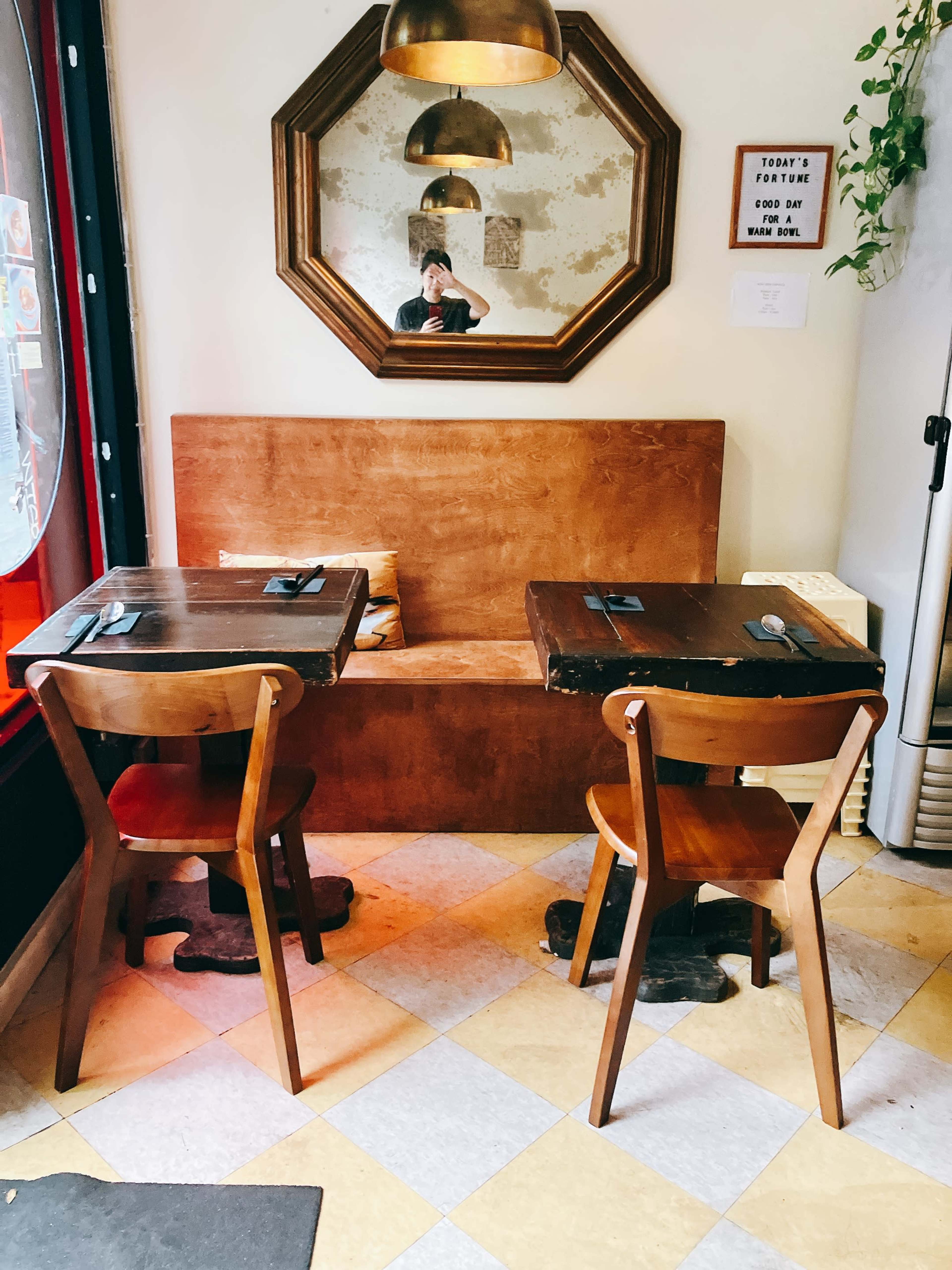 A cozy corner of a restaurant features two wooden tables with chairs, a large mirror above them, and potted greenery nearby.