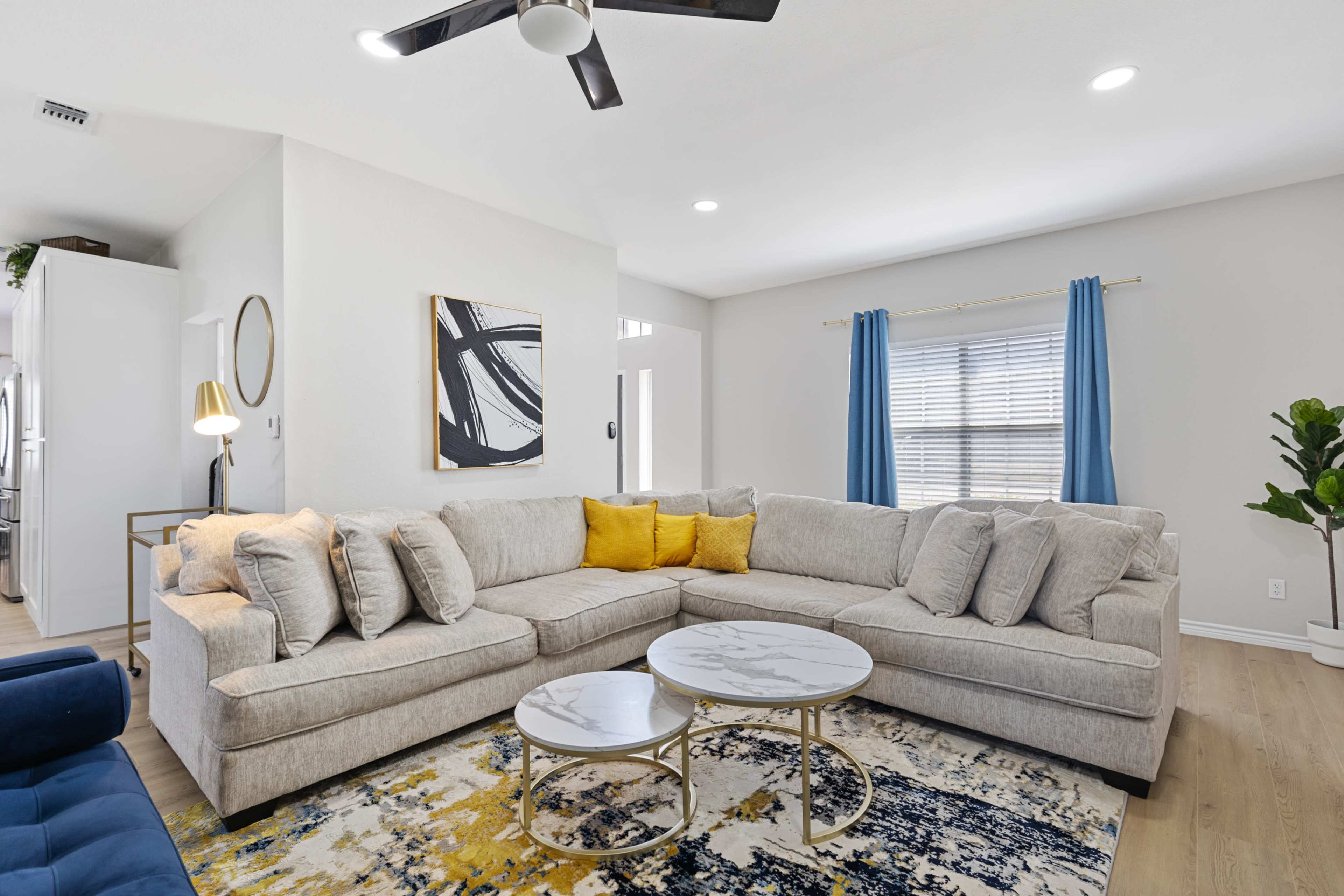 The image shows a modern living room featuring a light gray sectional sofa, yellow accent pillows, two round coffee tables, and a large area rug with blue and yellow patterns.