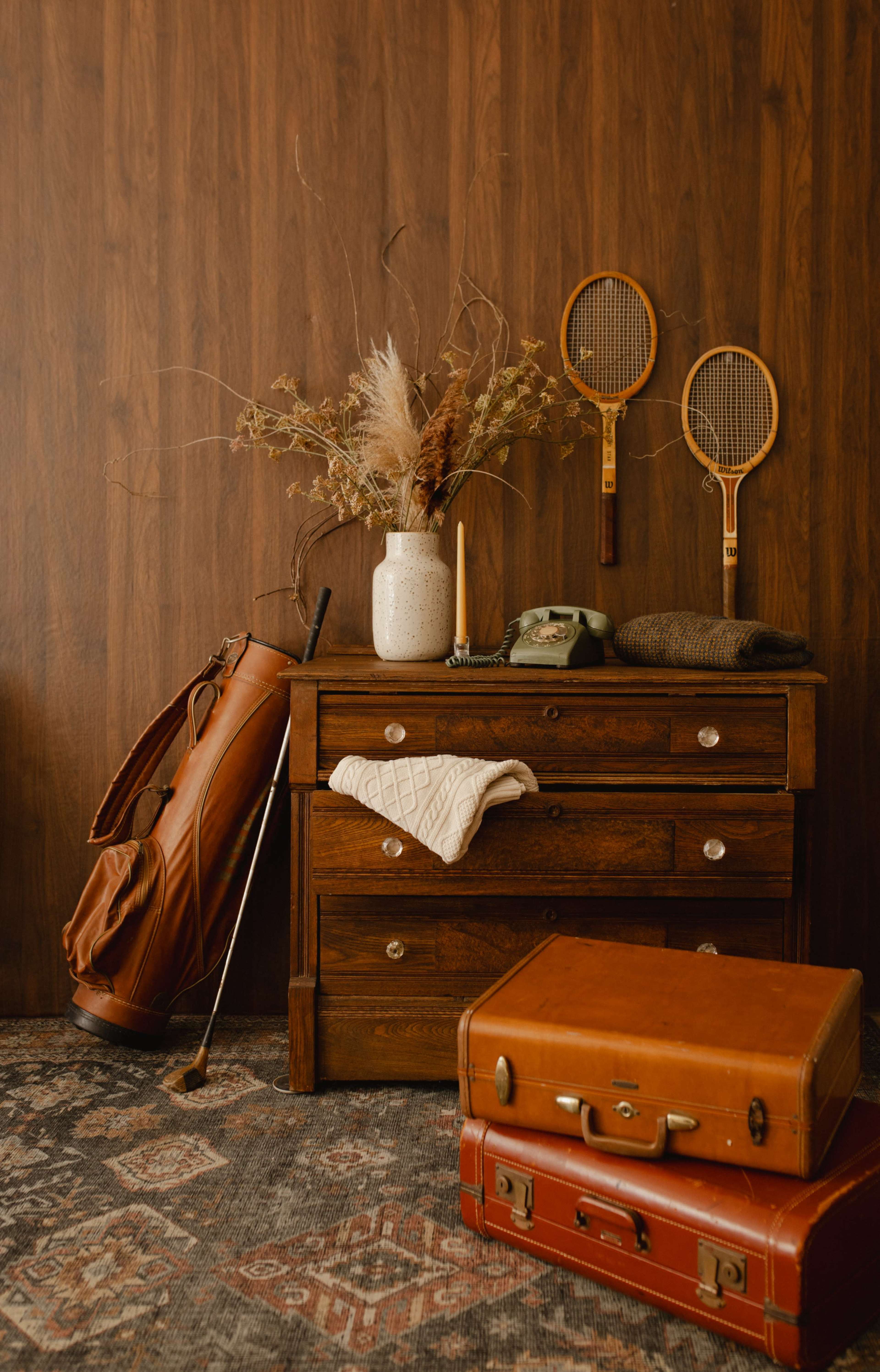 The image shows a wooden dresser with a textured rug underneath, accompanied by a golf bag, an old suitcase, a ceramic vase with dried flowers, and two tennis rackets hanging on the wall.