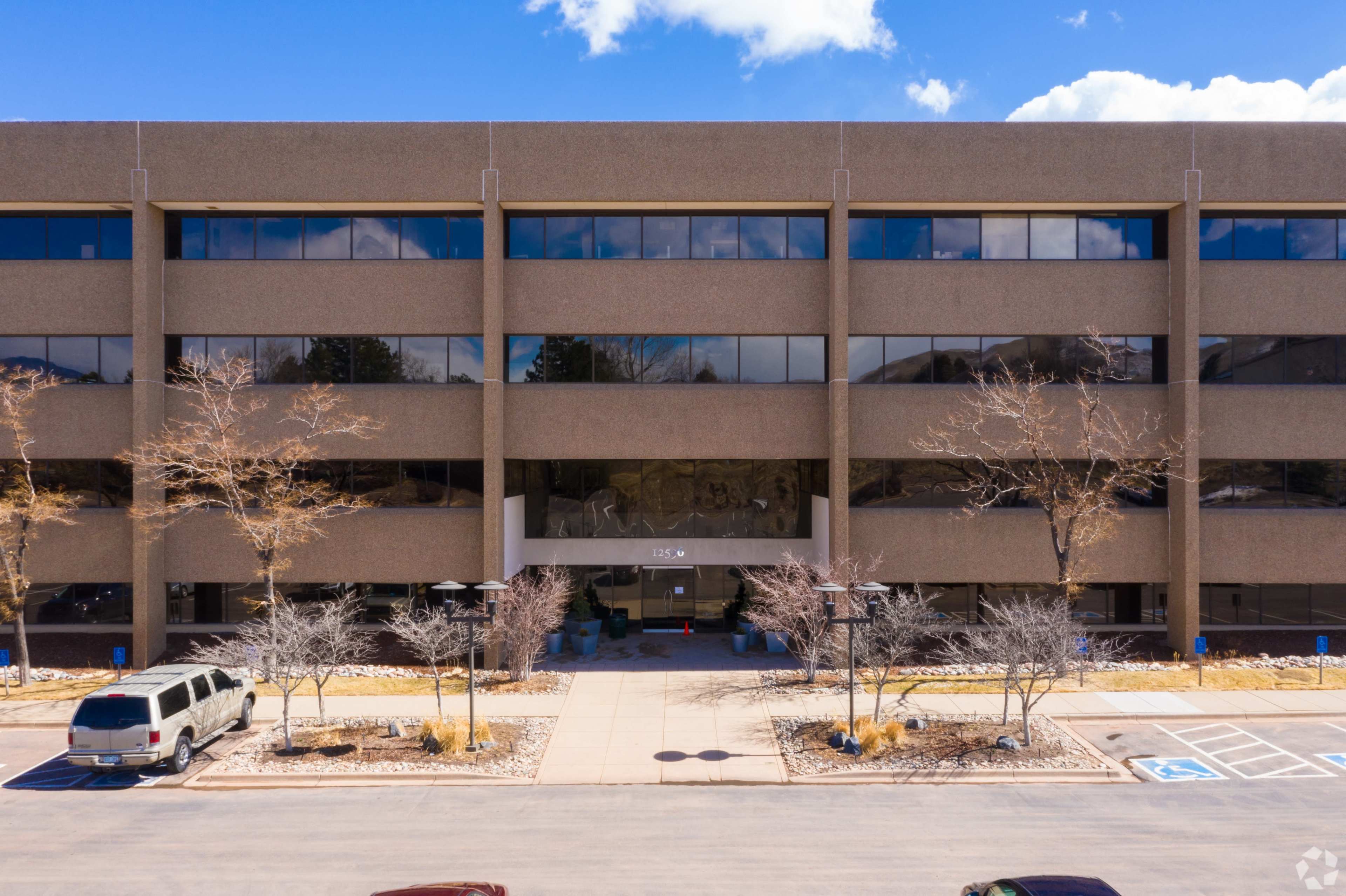 A four-story office building with a large glass entrance surrounded by paved walkways and parked cars.