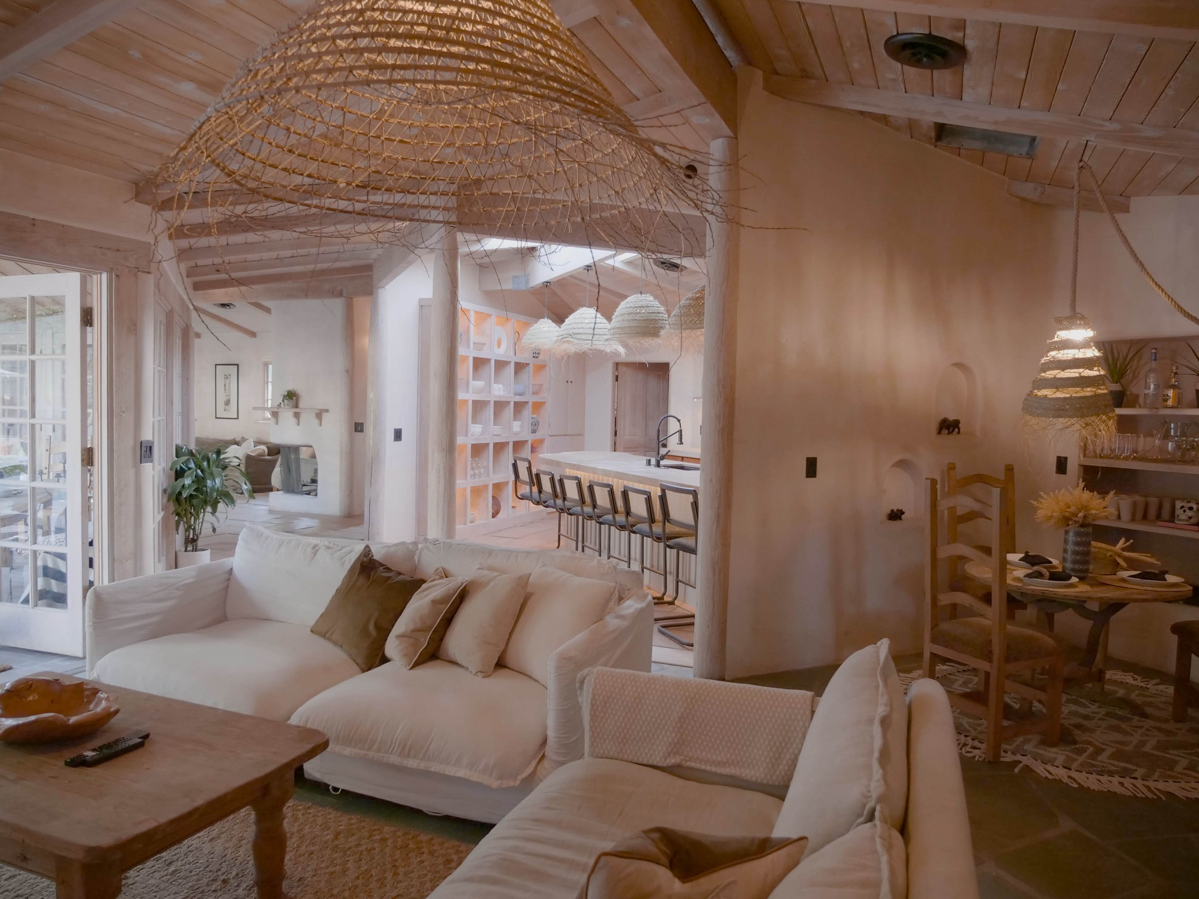 A spacious interior featuring a living area with a white sofa, a wooden coffee table, and a kitchen in the background with bar seating and hanging light fixtures.