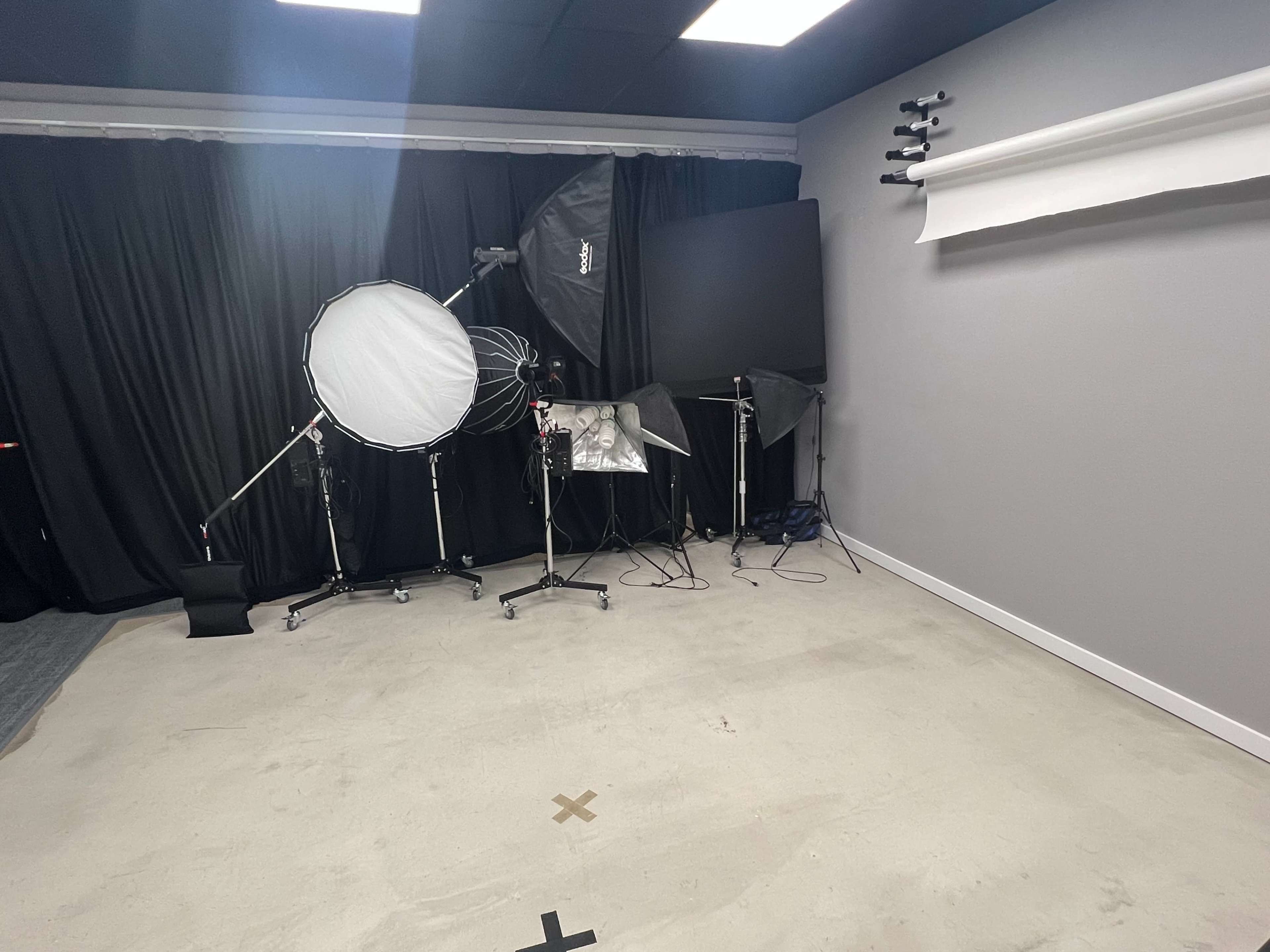 The image shows a photography studio featuring various lighting equipment, including a large softbox and a backdrop, with a plain floor and dark curtains.