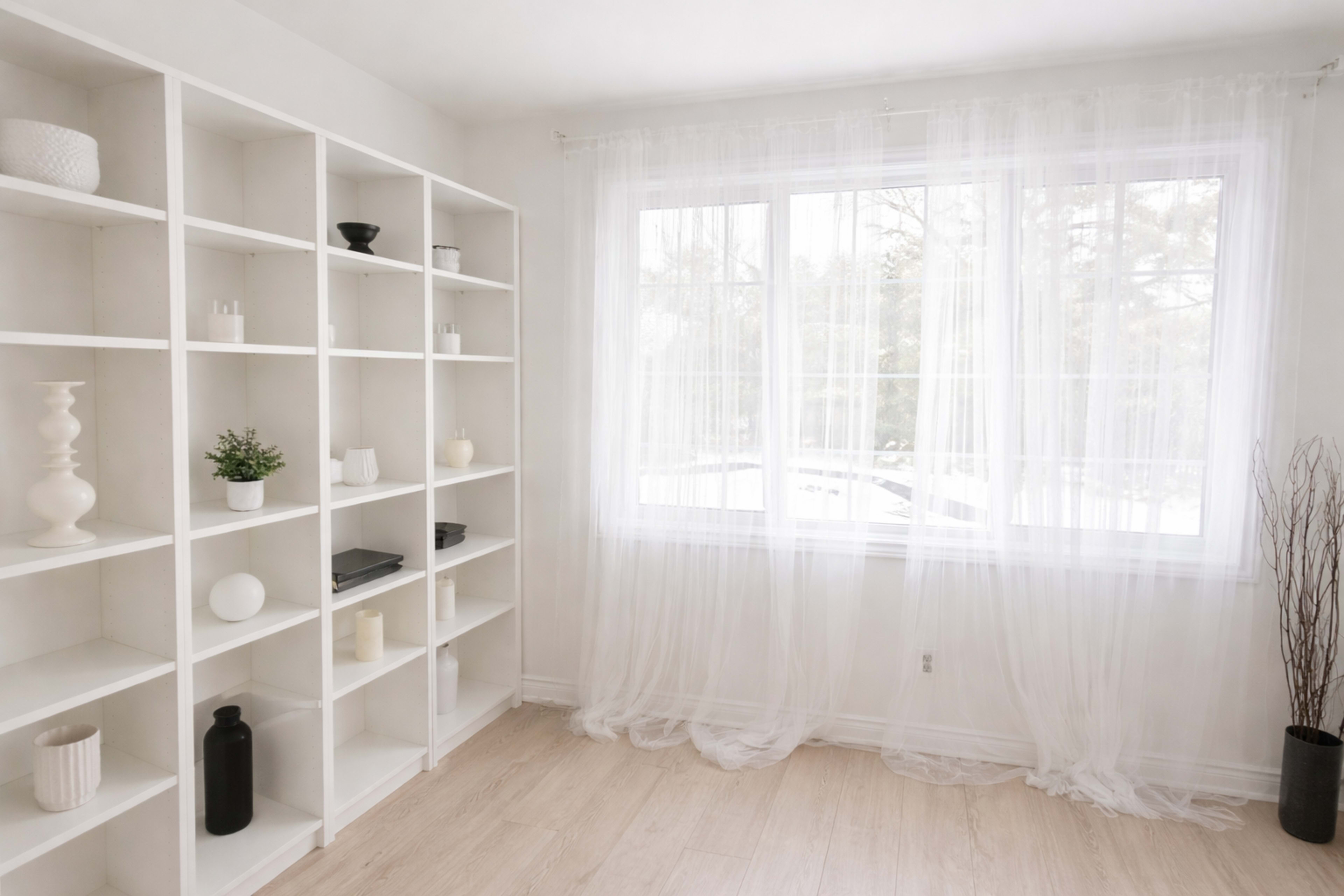 A bright room features a white shelving unit filled with decorative items and a large window draped with sheer curtains overlooking a snowy scene outside.
