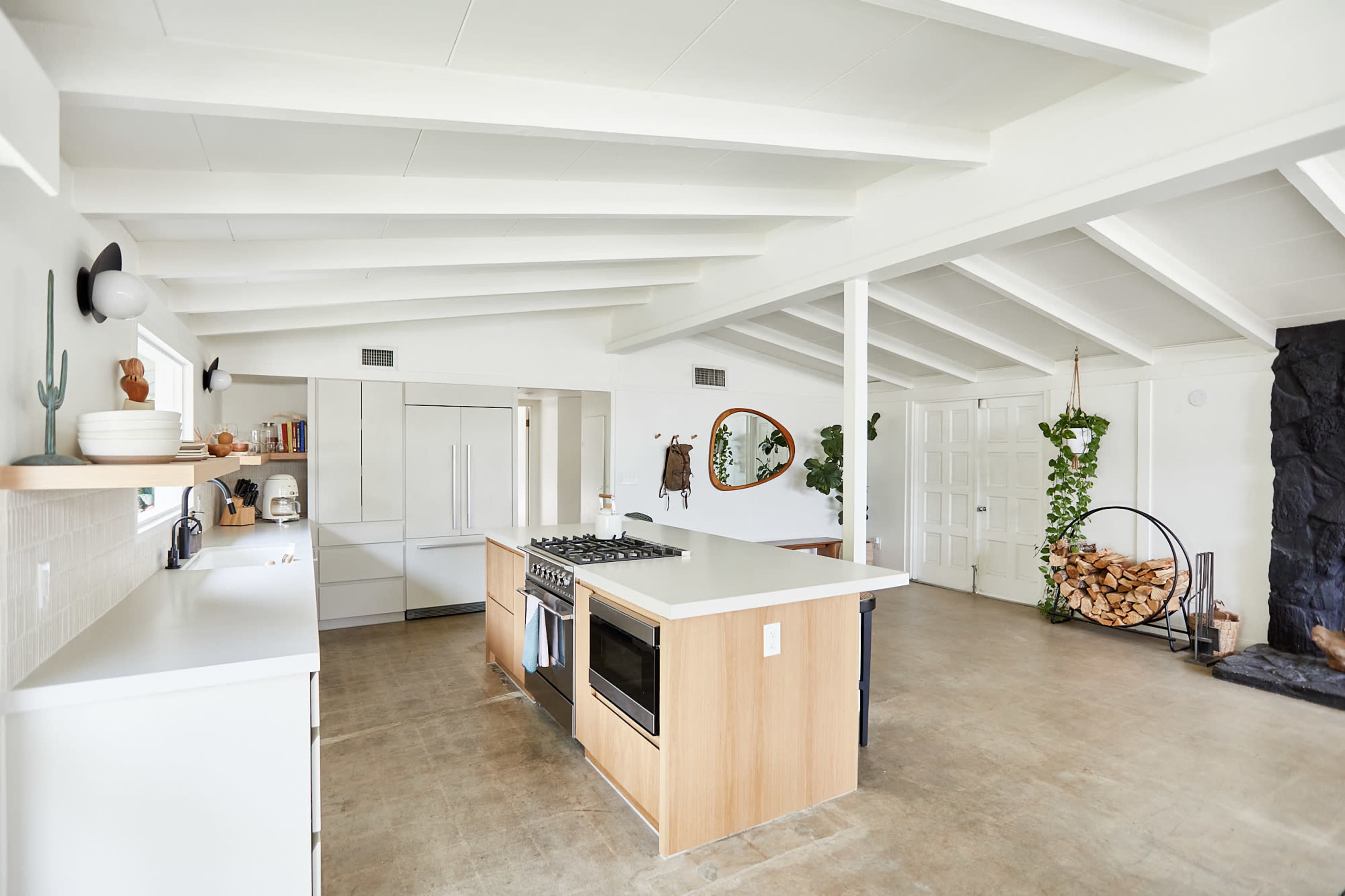 A modern kitchen features a large island with a gas stove, open shelving, and natural wood accents, set in a bright space with exposed beam ceilings.