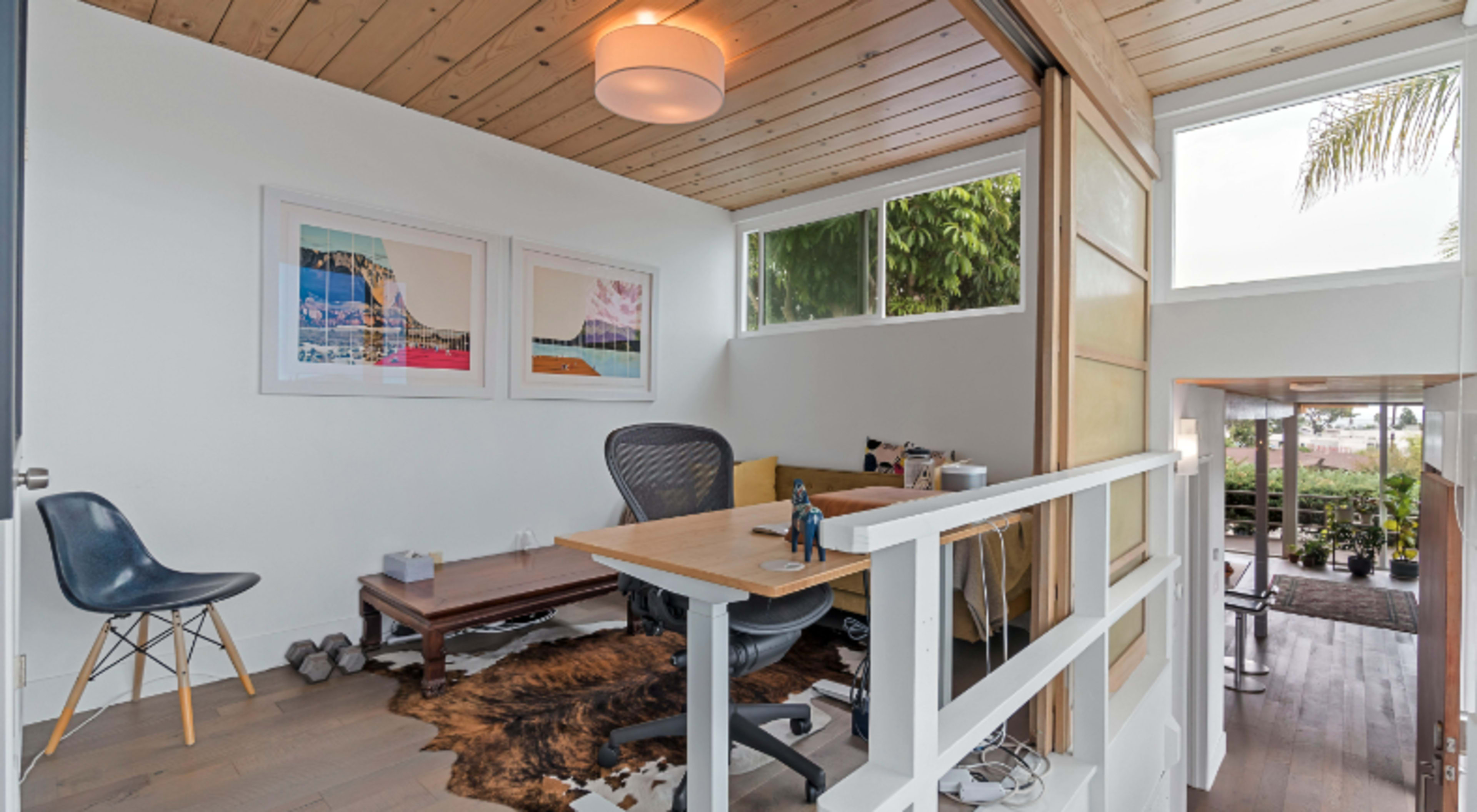 A modern home office with a wooden ceiling, a desk, an ergonomic chair, and framed artworks on the wall.