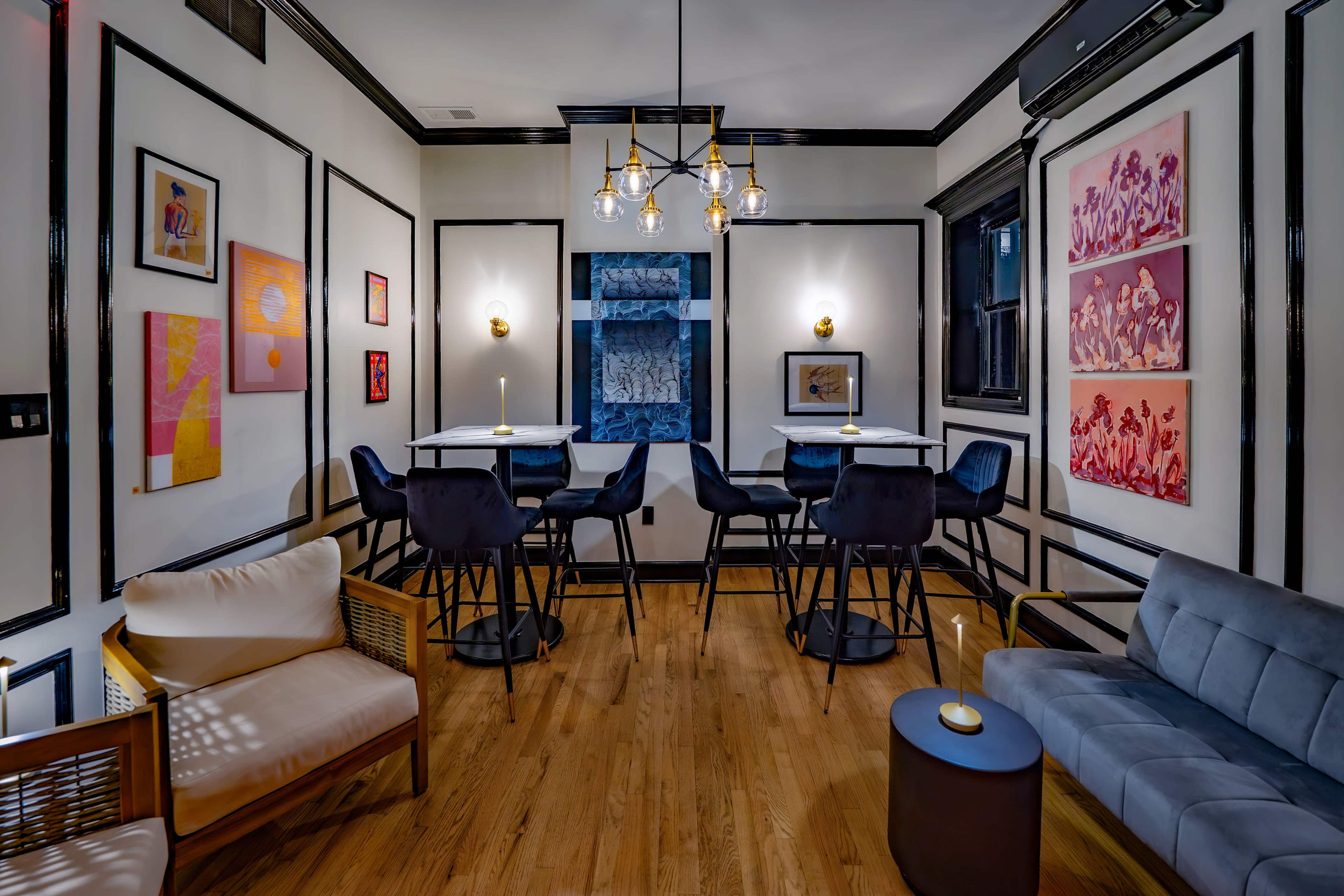 The image shows a modern interior with two seating areas, featuring a combination of high tables and low seating against a backdrop of framed artwork and stylish lighting.