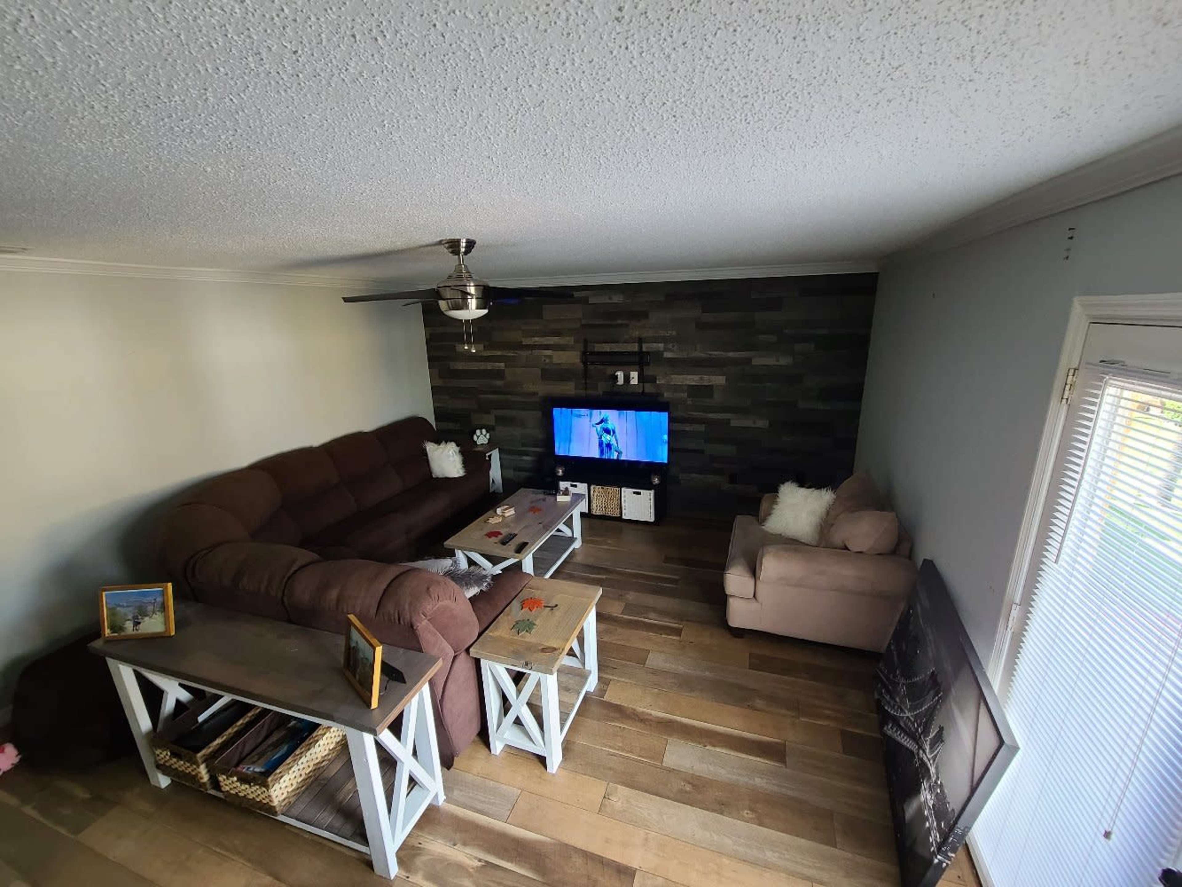 A living room features a brown sectional couch, two wooden tables, a television mounted on a stone accent wall, and a beige armchair.