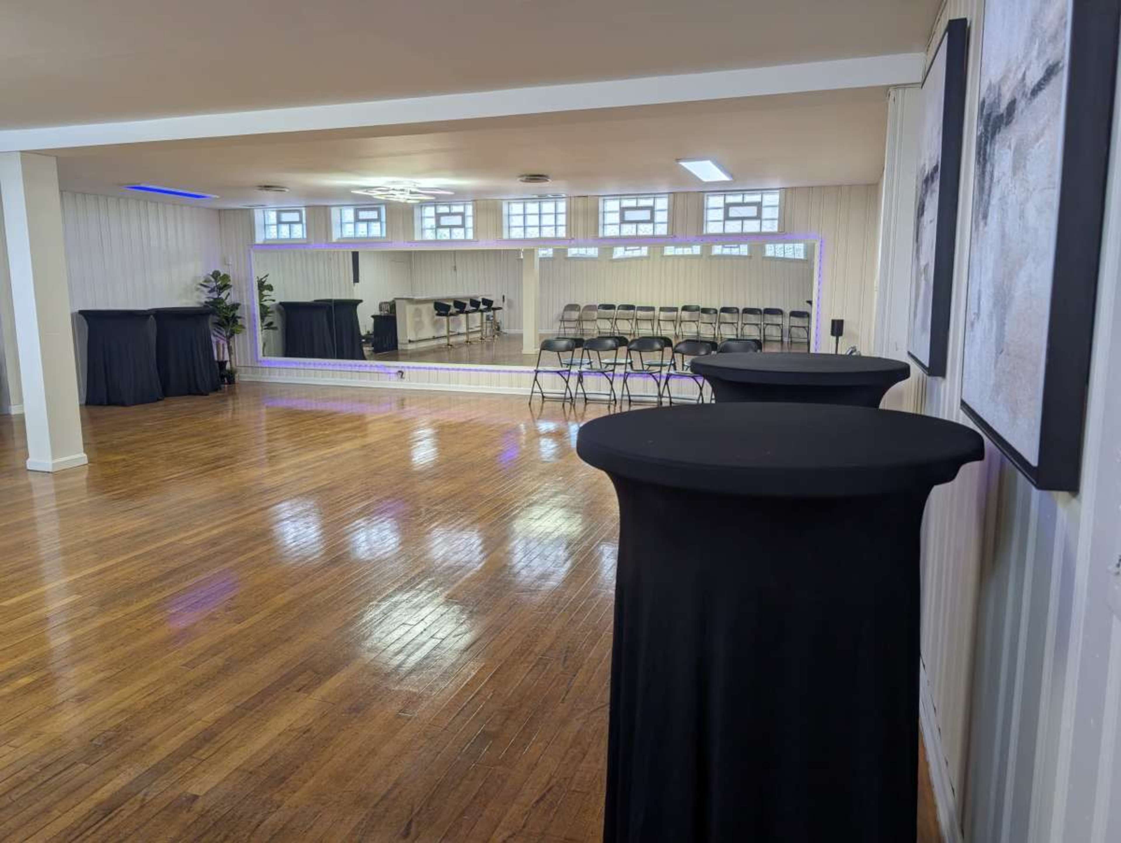 The image shows a spacious event room with polished wooden floors, black cocktail tables, and rows of chairs set up facing a mirrored wall.