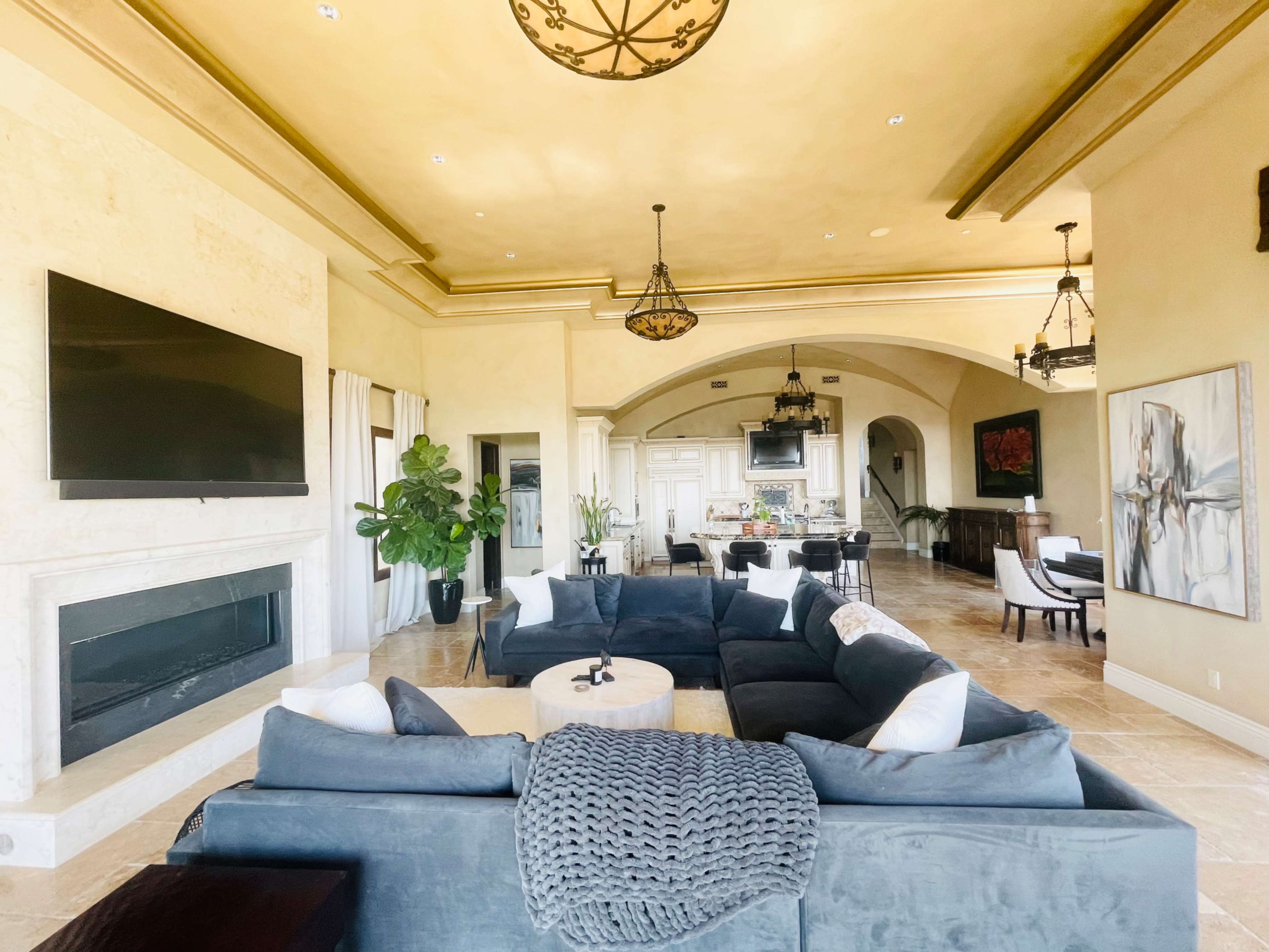 The living room features a large sectional sofa, a wall-mounted television above a fireplace, and an open layout leading to a kitchen area.