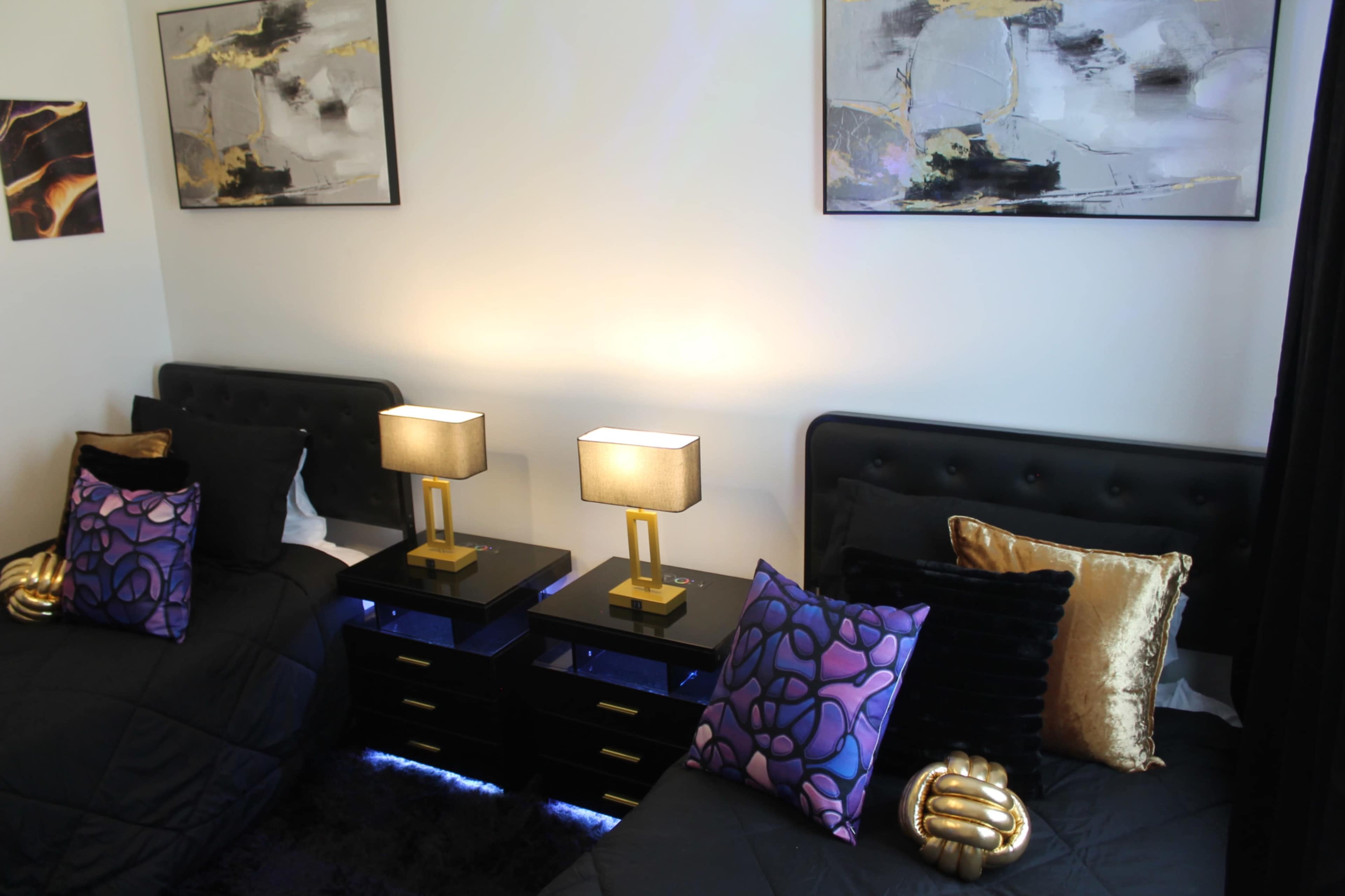 A modern shared bedroom with two black beds, each accompanied by a gold lamp and decorative pillows, against a backdrop of abstract art on the walls.